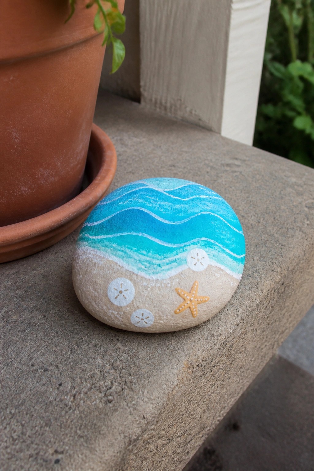 Painted rock with blue waves, sandy beach, white shells, and orange starfish on ledge