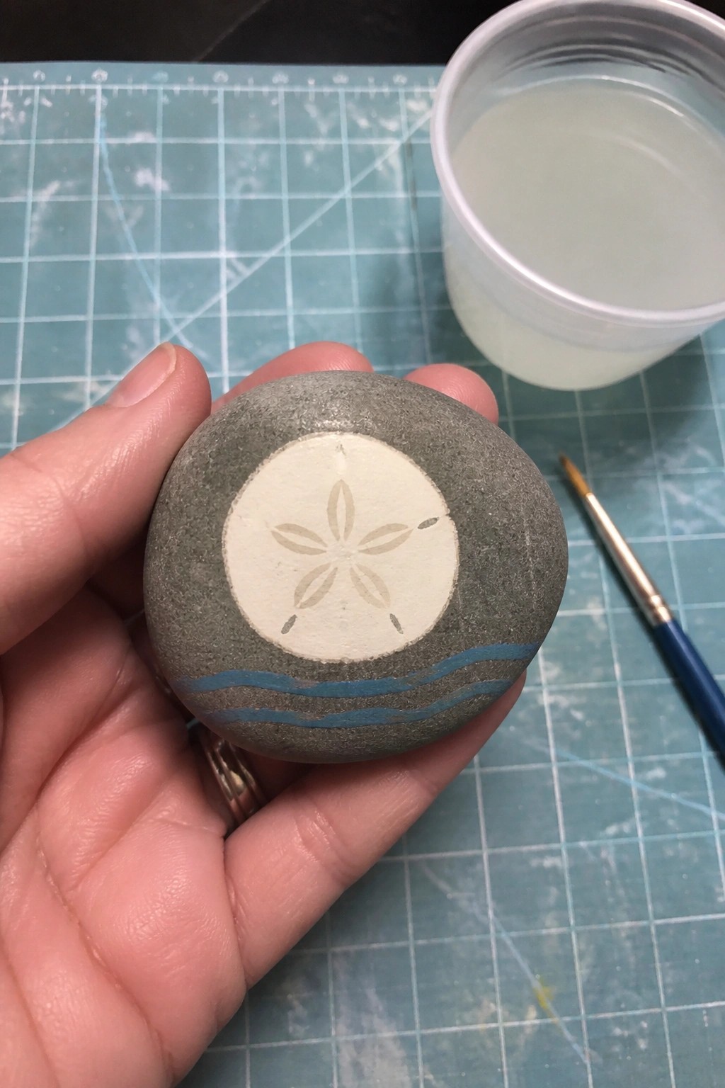 A hand holds a gray rounded rock painted with a sand dollar design and blue waves, next to a paintbrush and cup on a cutting mat.