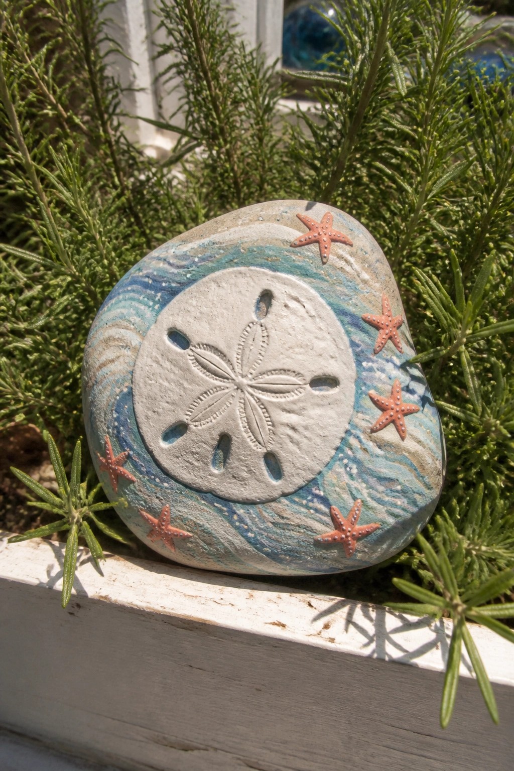 Painted stone with sand dollar and orange starfish amid green plants on white ledge