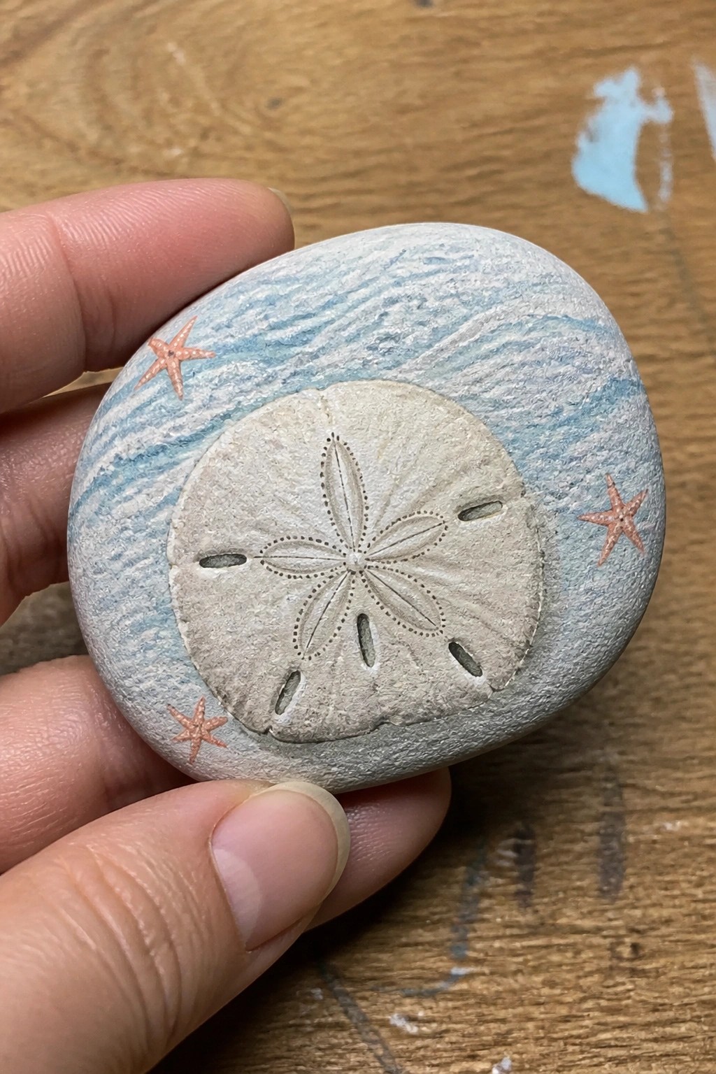 Hand holding painted rock with central sand dollar and orange starfish on blue waves.