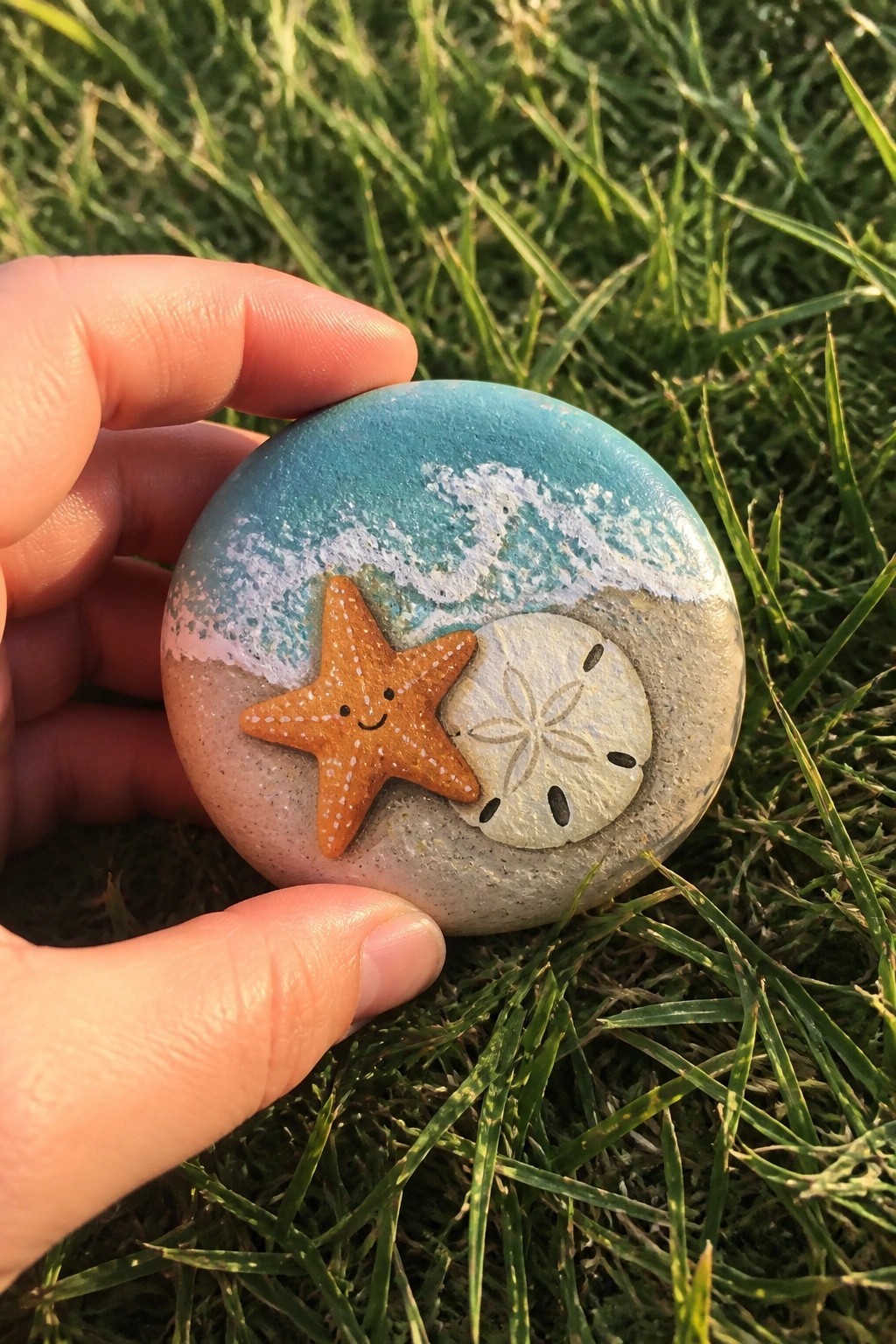 Hand holding round rock painted with ocean wave, smiling starfish, and sand dollar