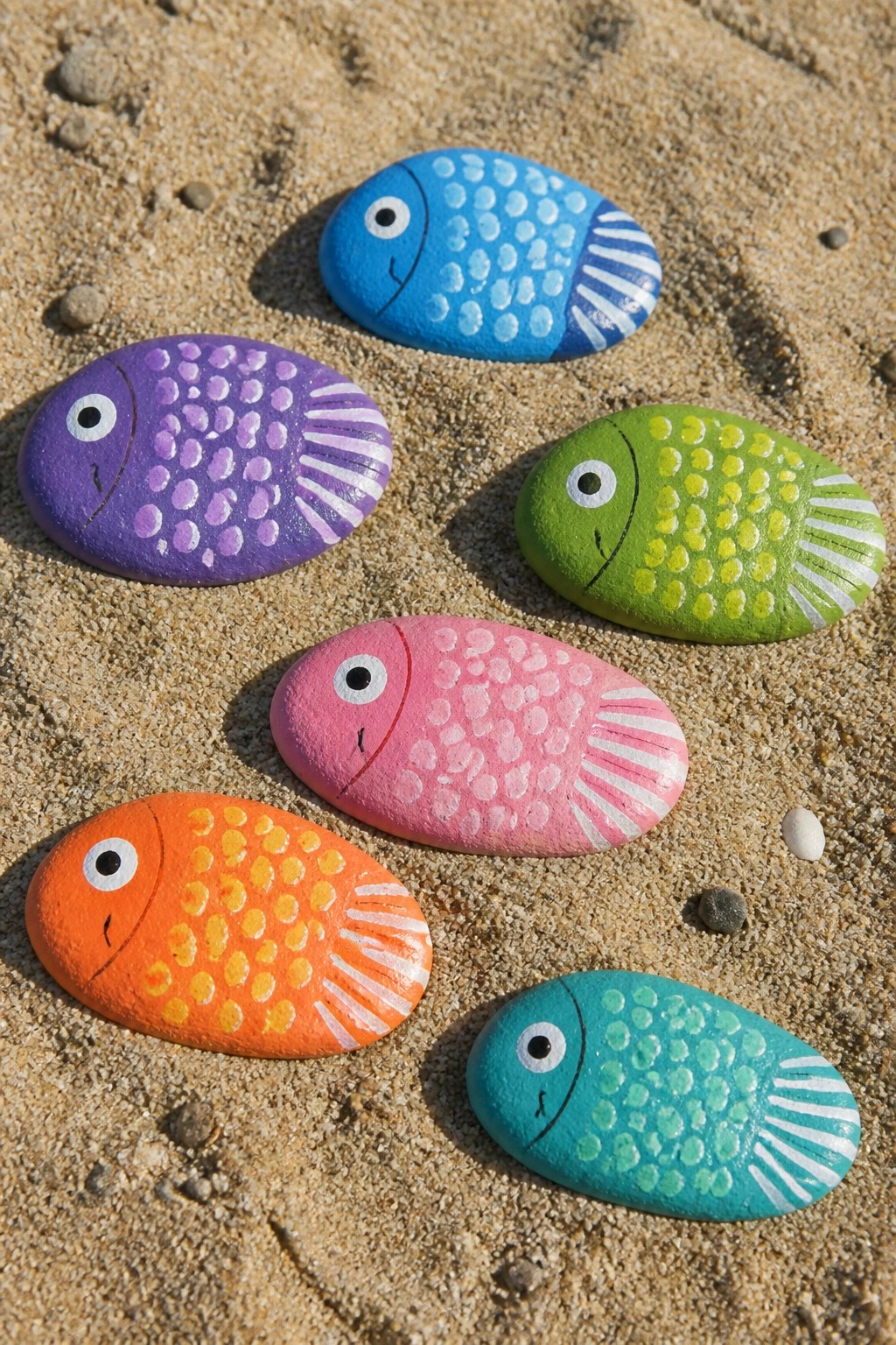 Colorful painted fish rocks in blue, purple, green, pink, orange, and teal on sand.