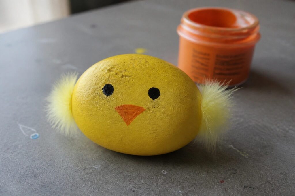 22 Easy Painted Rocks for Kids to Make at Camps, Classrooms, and Home Yellow egg chick with black eyes, orange beak, and yellow feathers on gray table
