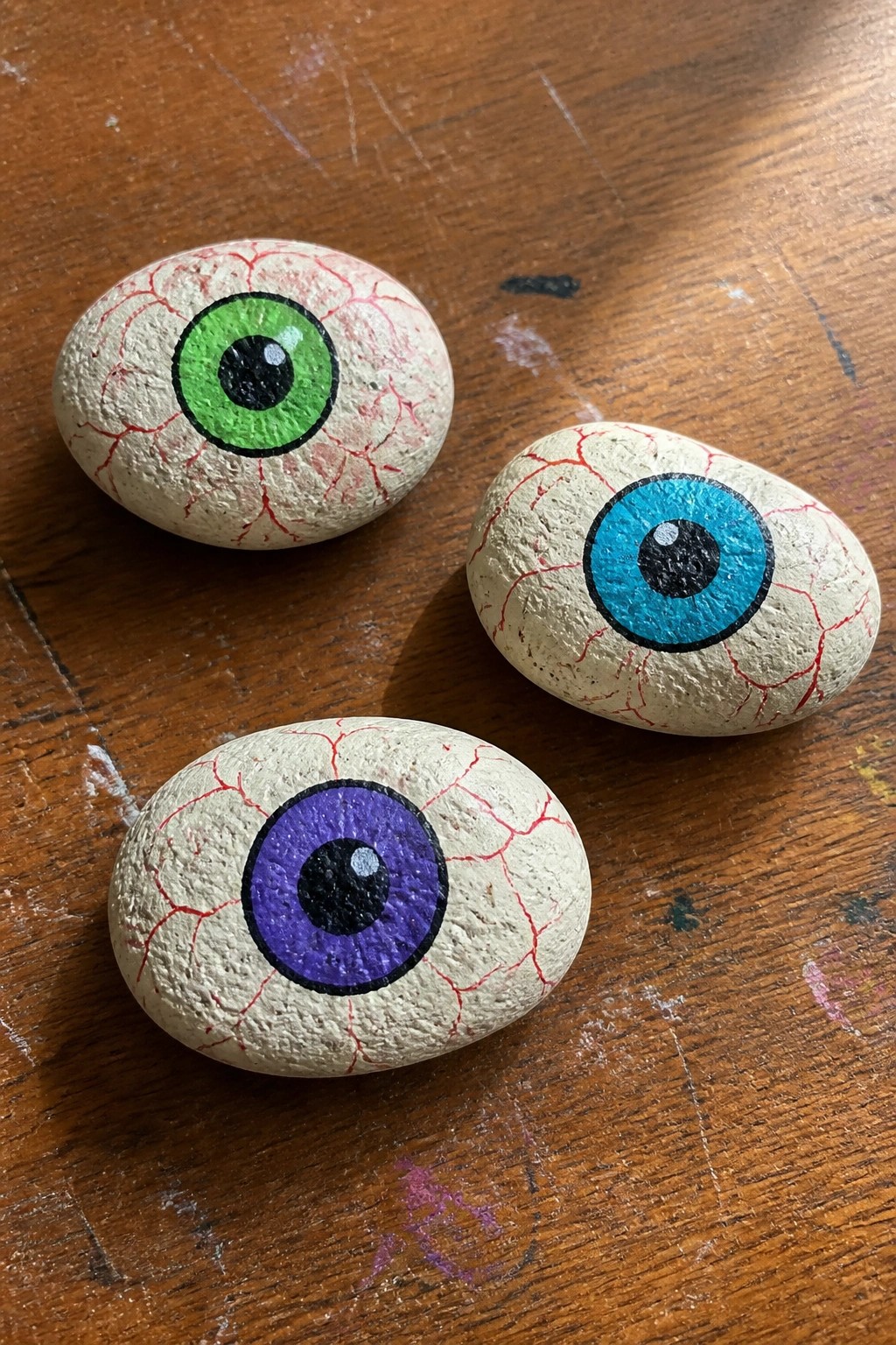Three rounded rocks painted as eyeballs with red veins, green blue and purple irises, and black pupils rest on a wooden table.