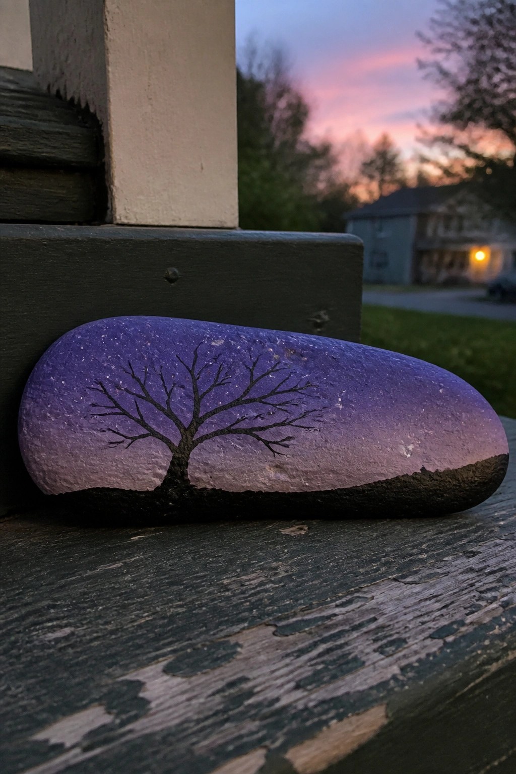 Painted rock with black tree silhouette on purple starry gradient sky atop wood.