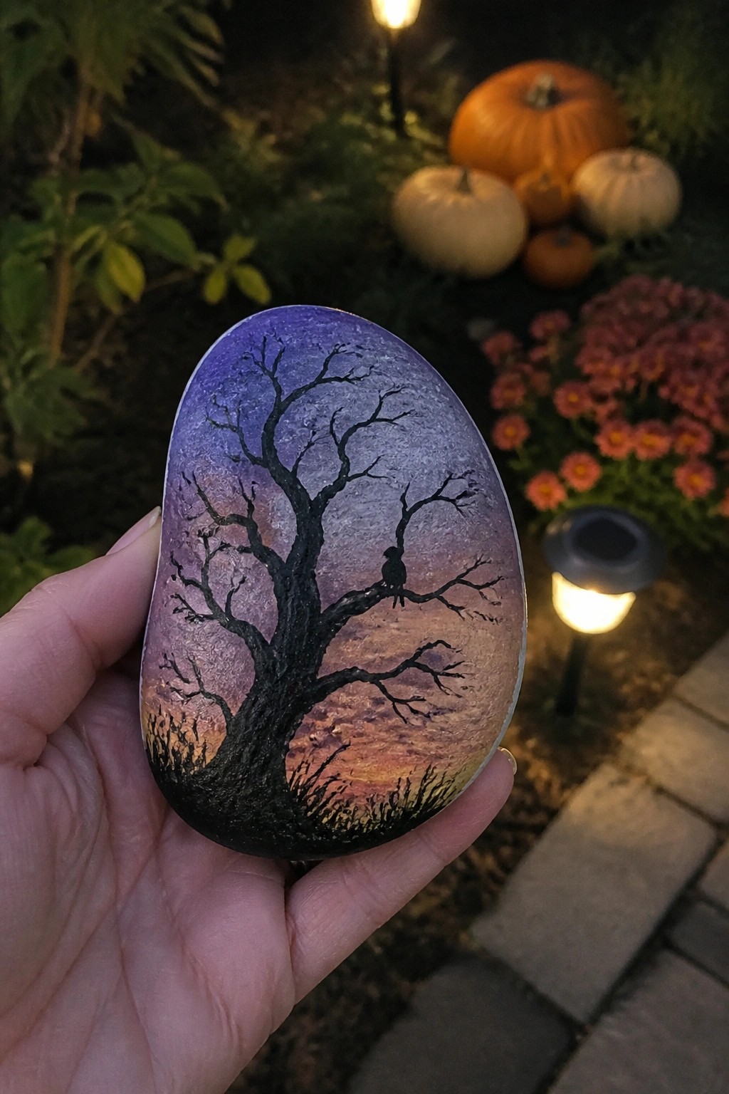 Hand holding painted stone with bare tree, bird, and purple-orange sunset sky.
