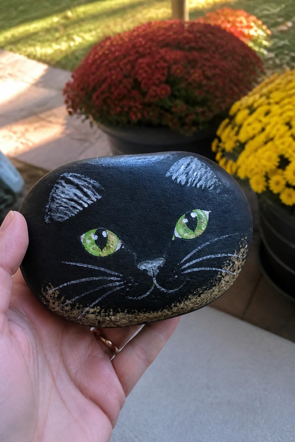 A hand holds a smooth black painted rock with a green-eyed cat face and white whiskers in front of red and yellow flower pots.