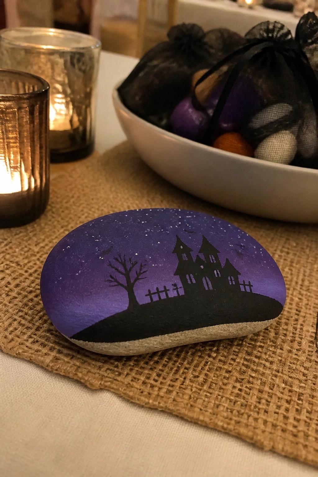 A smooth oval stone painted with a purple and black night sky, black haunted house silhouette, tree, fence, and bats sits on burlap near candles.