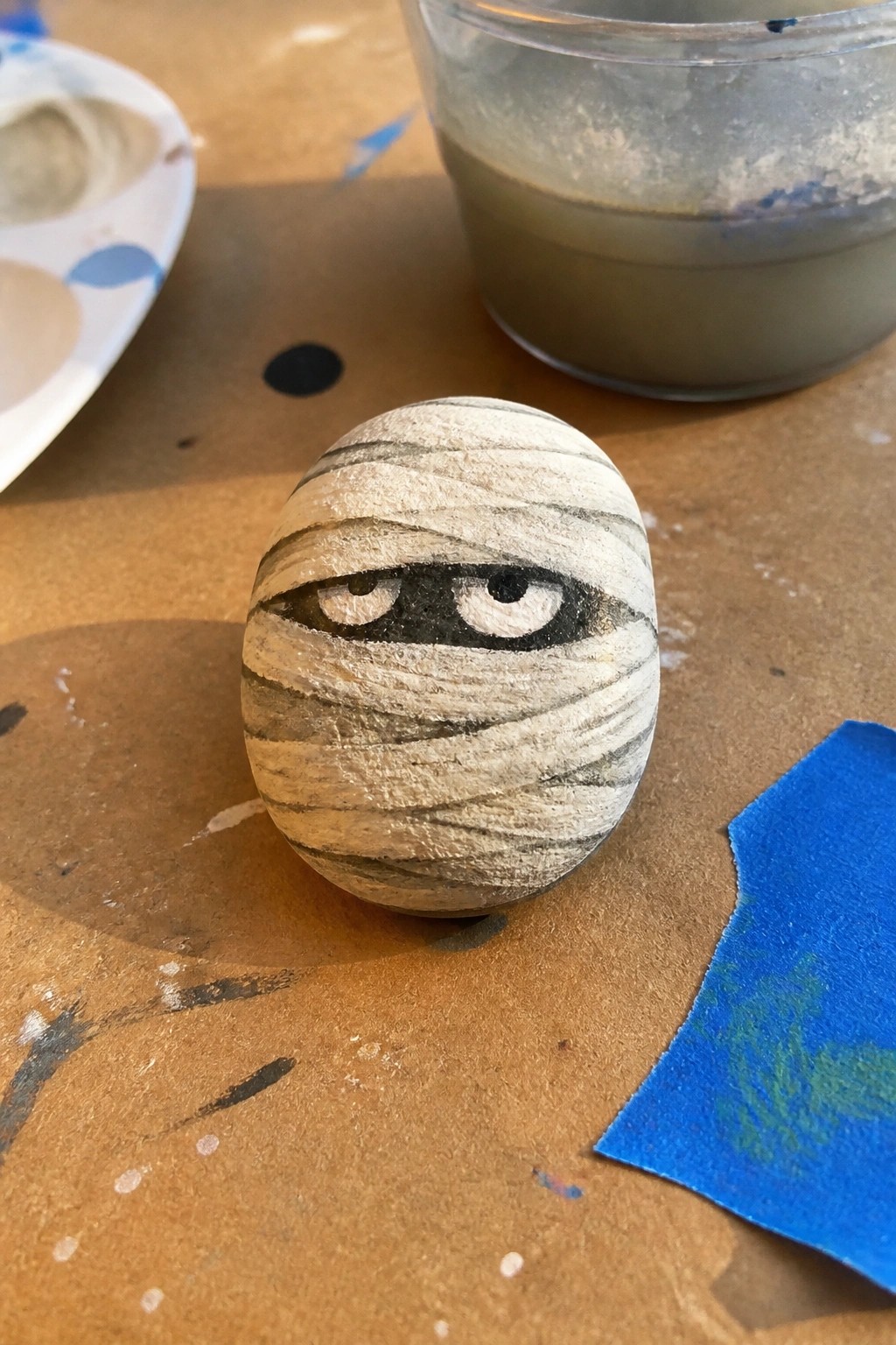 Painted egg mummy with striped bandages and cartoon eyes amid art supplies