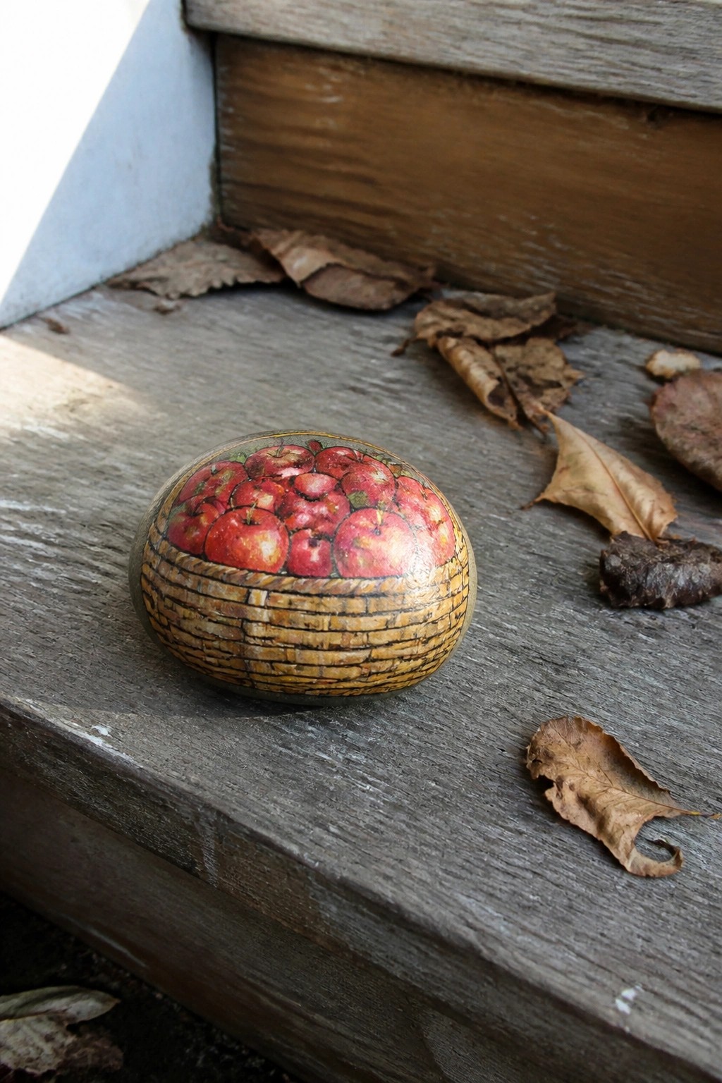 Painted stone with basket of red apples on wooden steps amid dry leaves