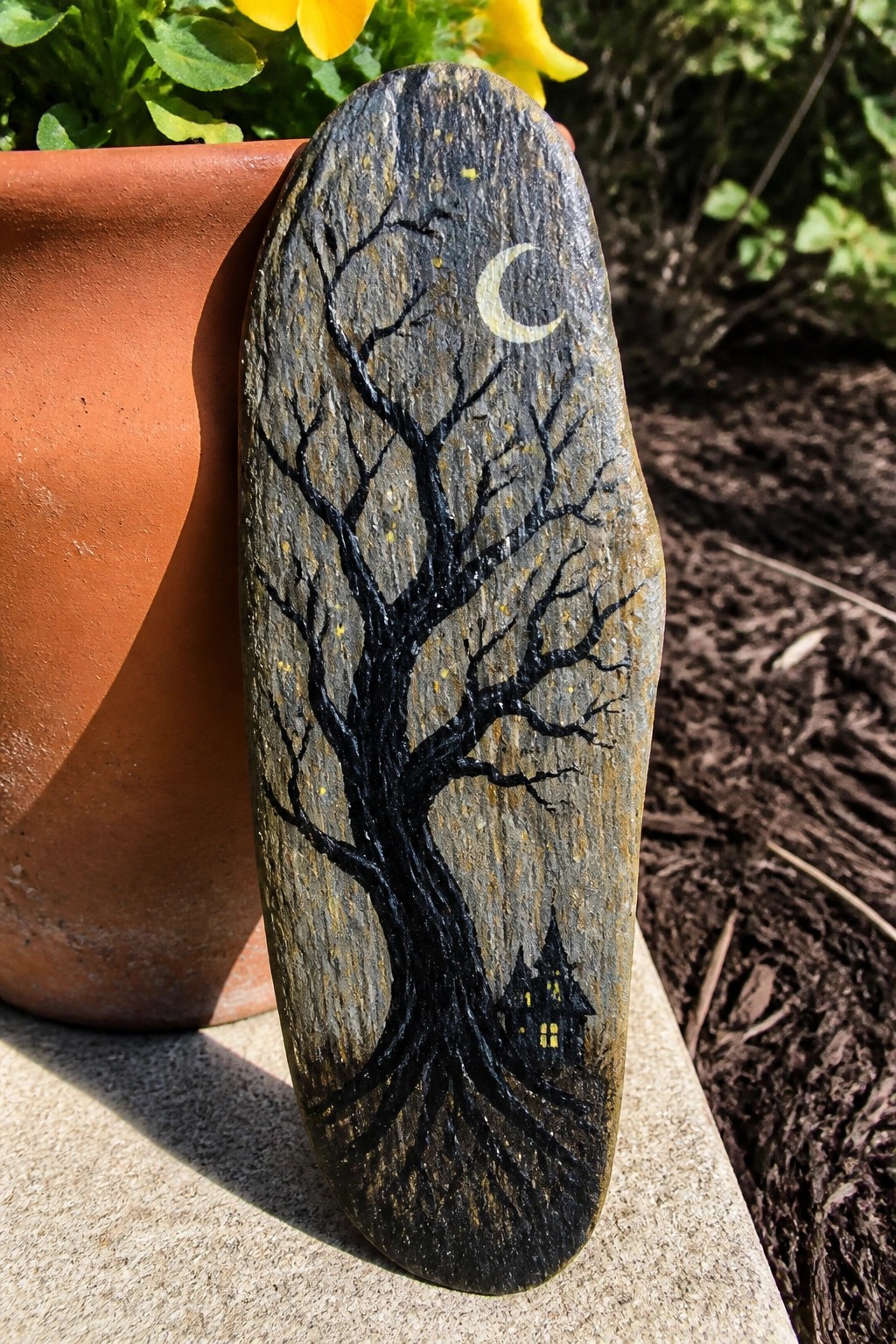 A painted rock shows a black tree silhouette with a crescent moon above and a small house with lit windows at the base, leaning against a terracotta pot.