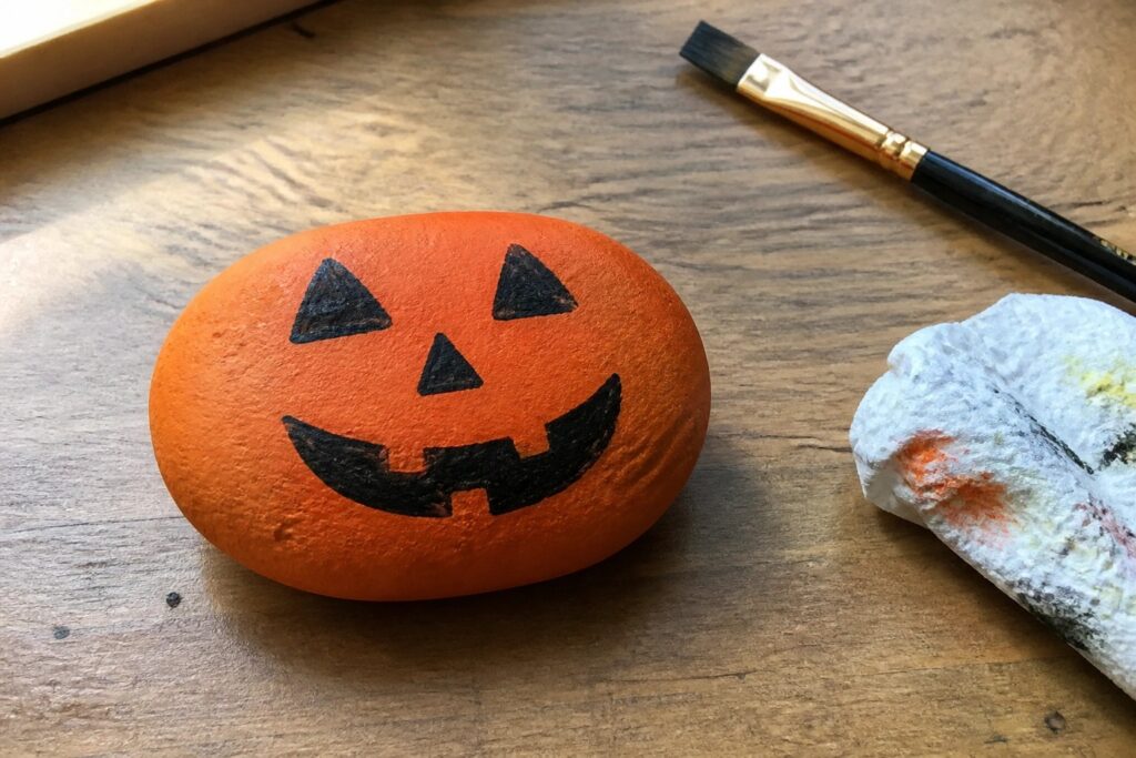 23 Budget-Friendly Halloween Rock Crafts to Make Decorations Without Expensive Supplies Orange rock painted as jack-o'-lantern face with paintbrush and stained towel on wood