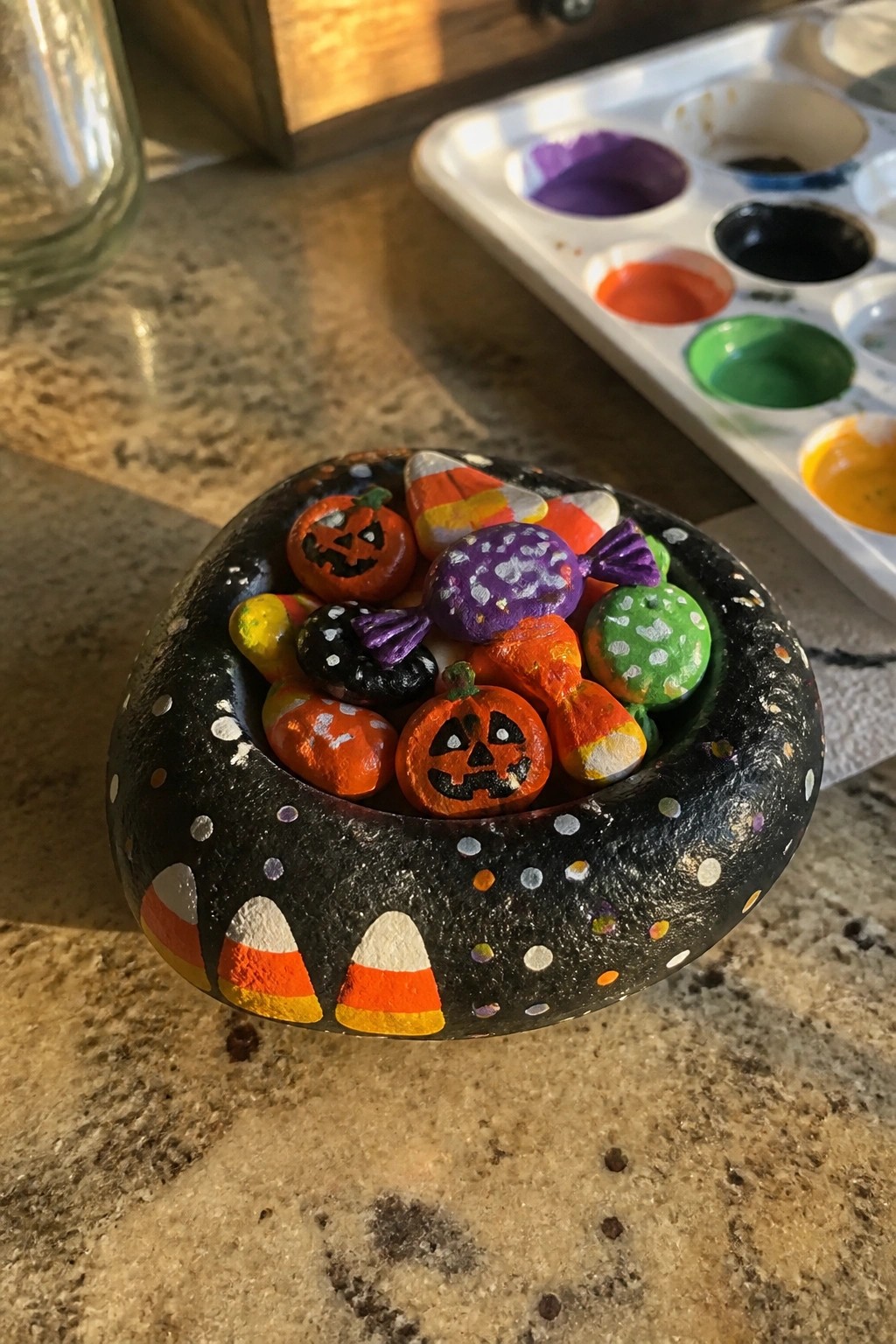 Black painted rock bowl with candy corn holding Halloween-themed painted rocks beside palette