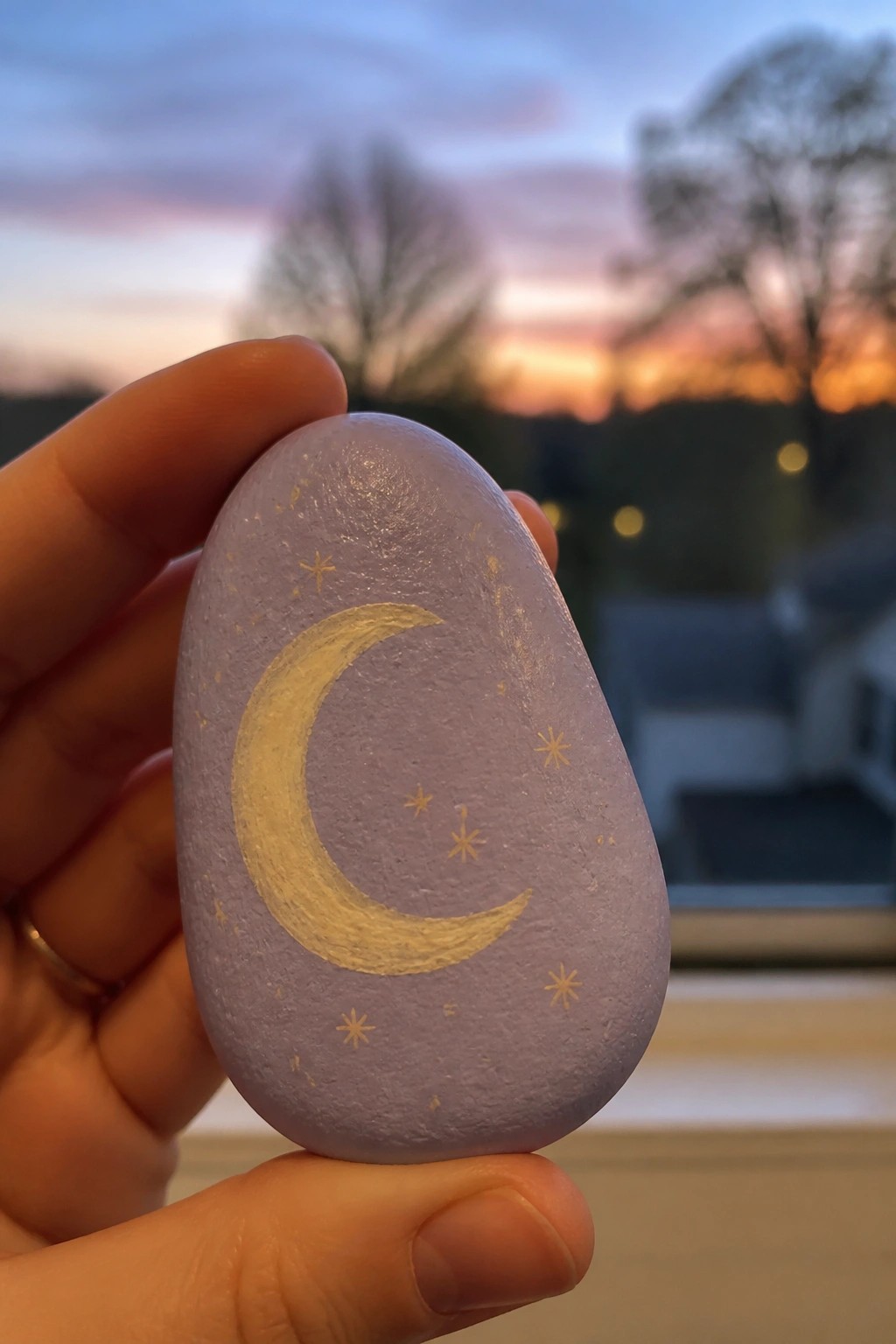 A hand holds a light purple painted rock with a gold crescent moon and scattered gold stars.