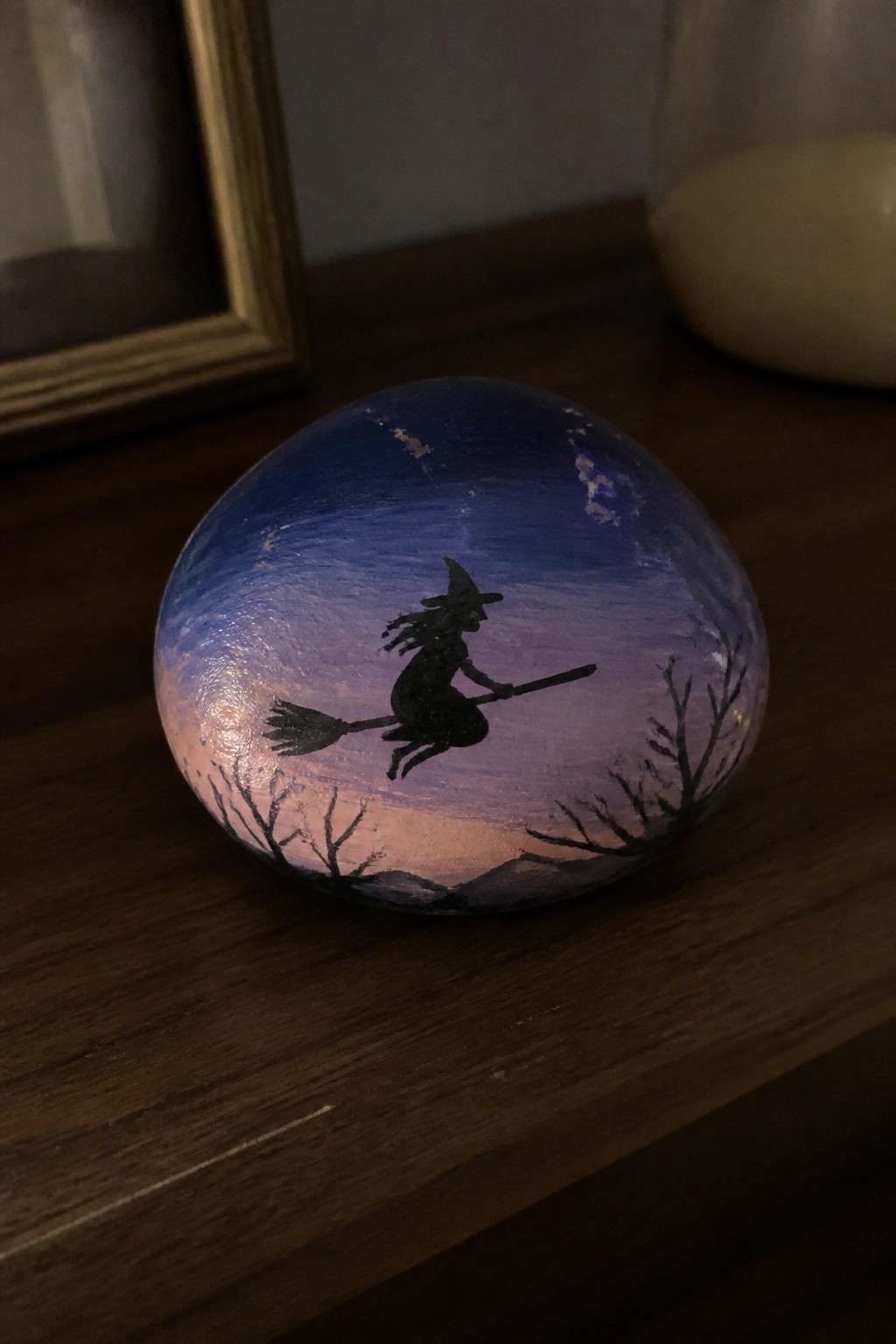 A round rock painted with a blue-purple night sky shows a black witch on a broomstick silhouette above bare tree branches.