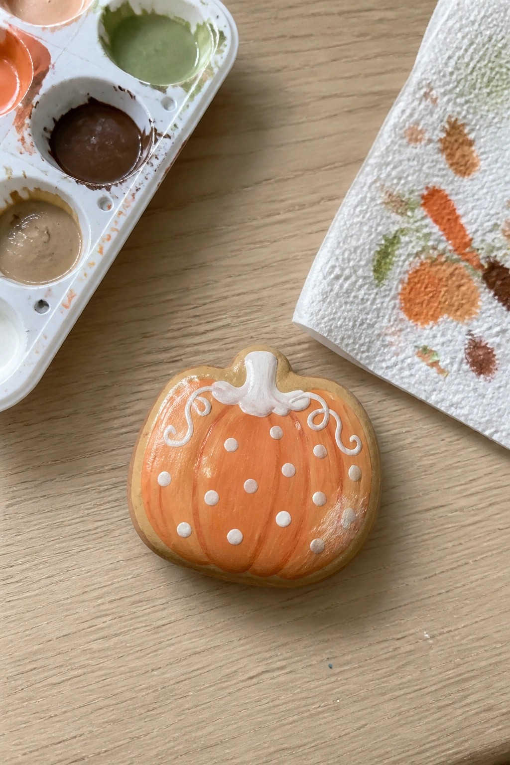 Orange pumpkin cookie with white dots and swirls beside watercolor palette on wood