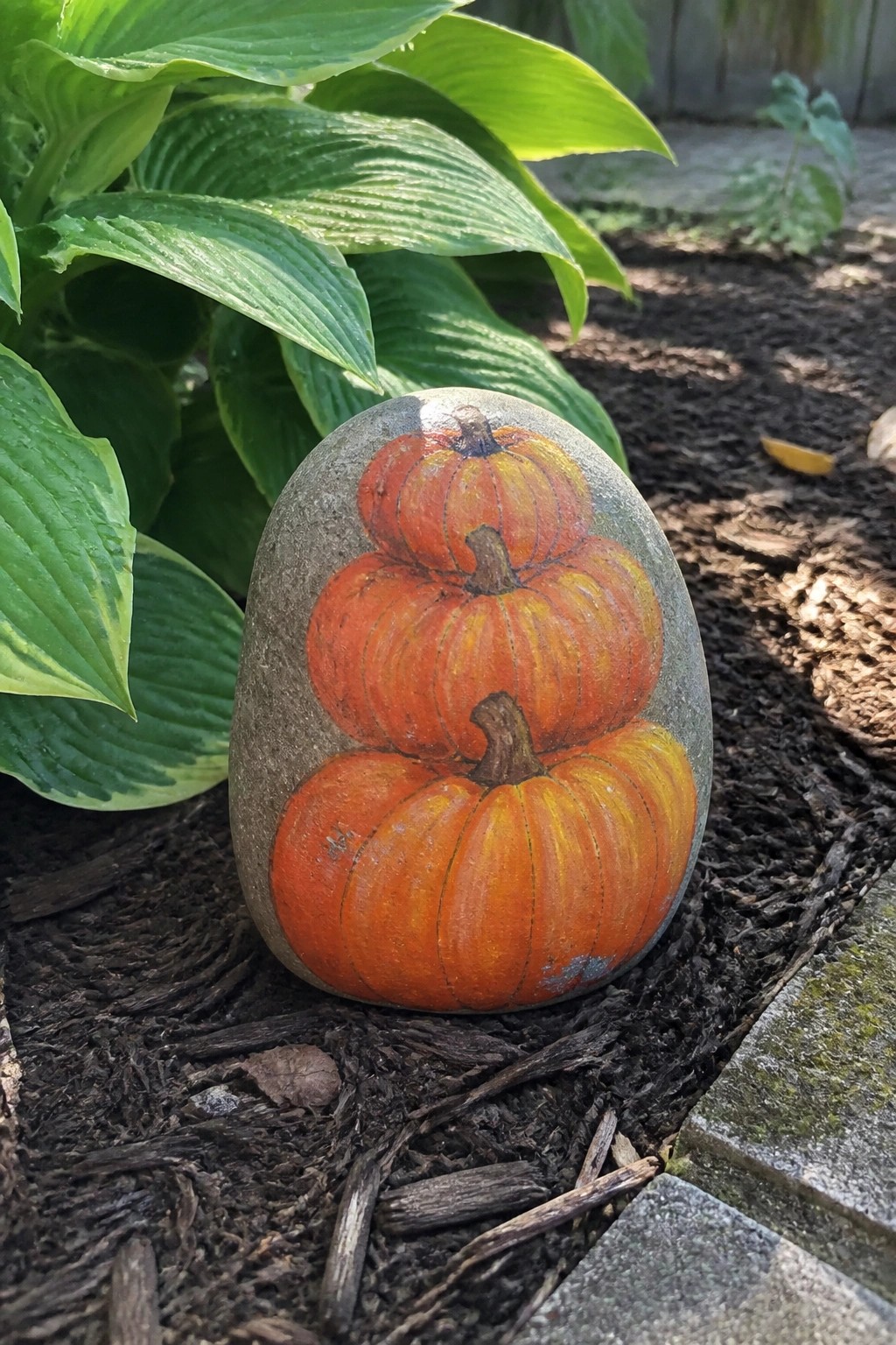 Painted rock with three stacked orange pumpkins on garden mulch beside green leaves.