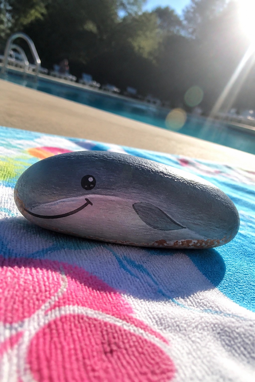 Smiling whale painted on gray rock atop colorful towel beside sunny pool