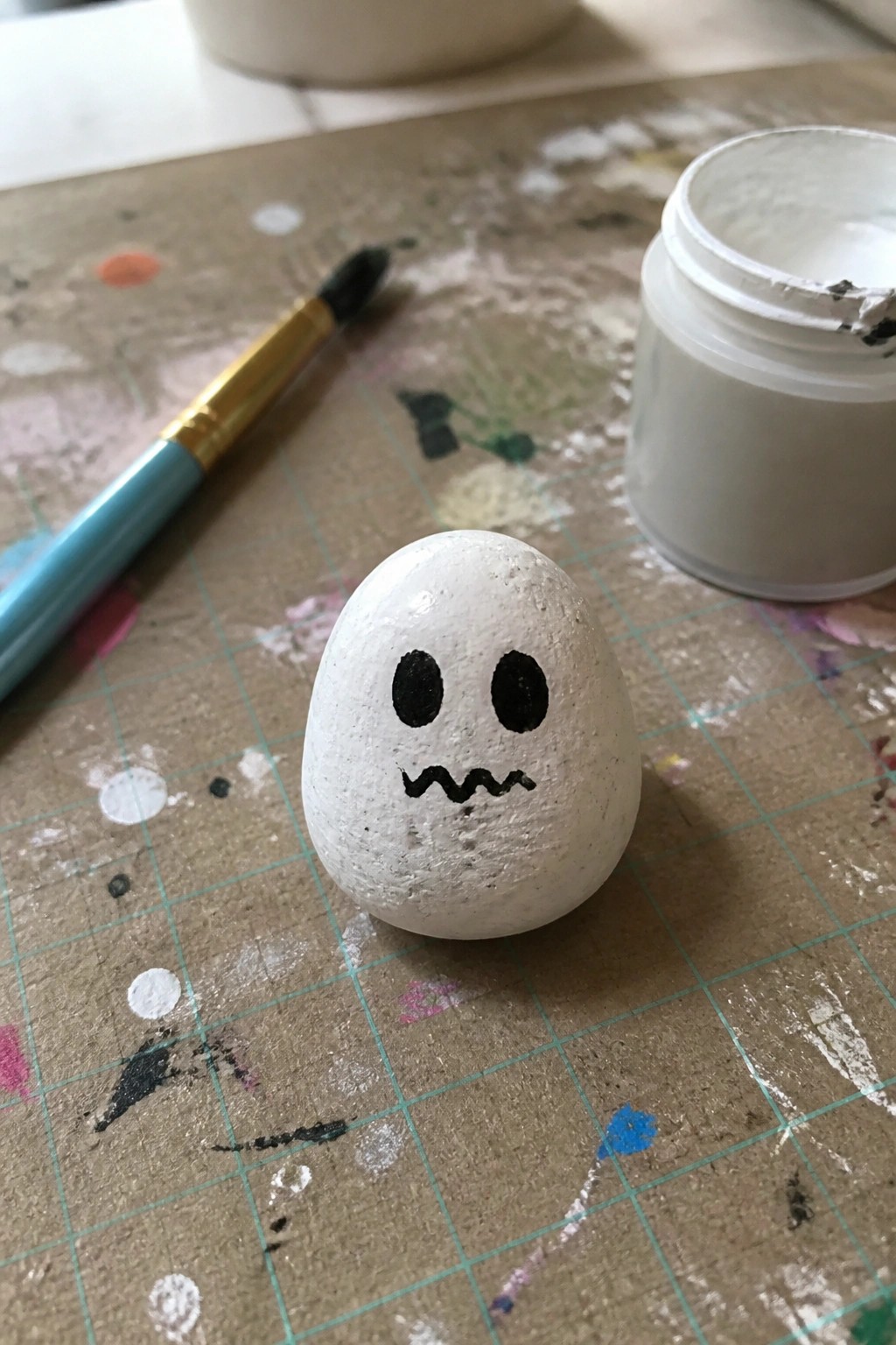 Painted ghost rock on splattered mat beside paintbrush and white paint jar