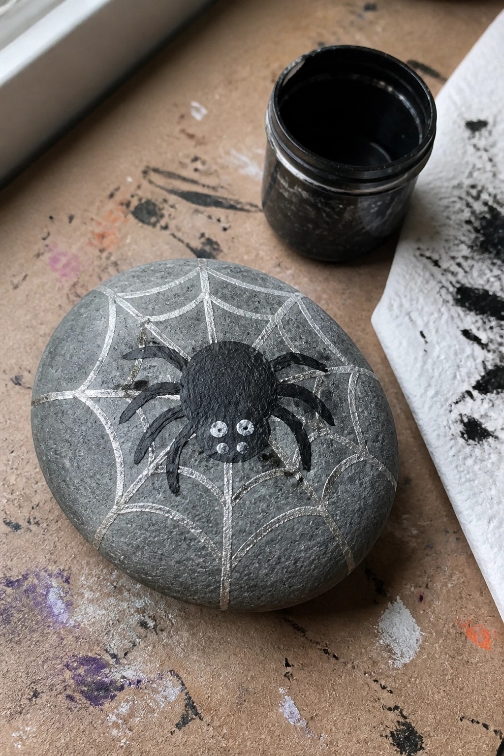 Painted gray rock with black spider on silver web beside paint jar