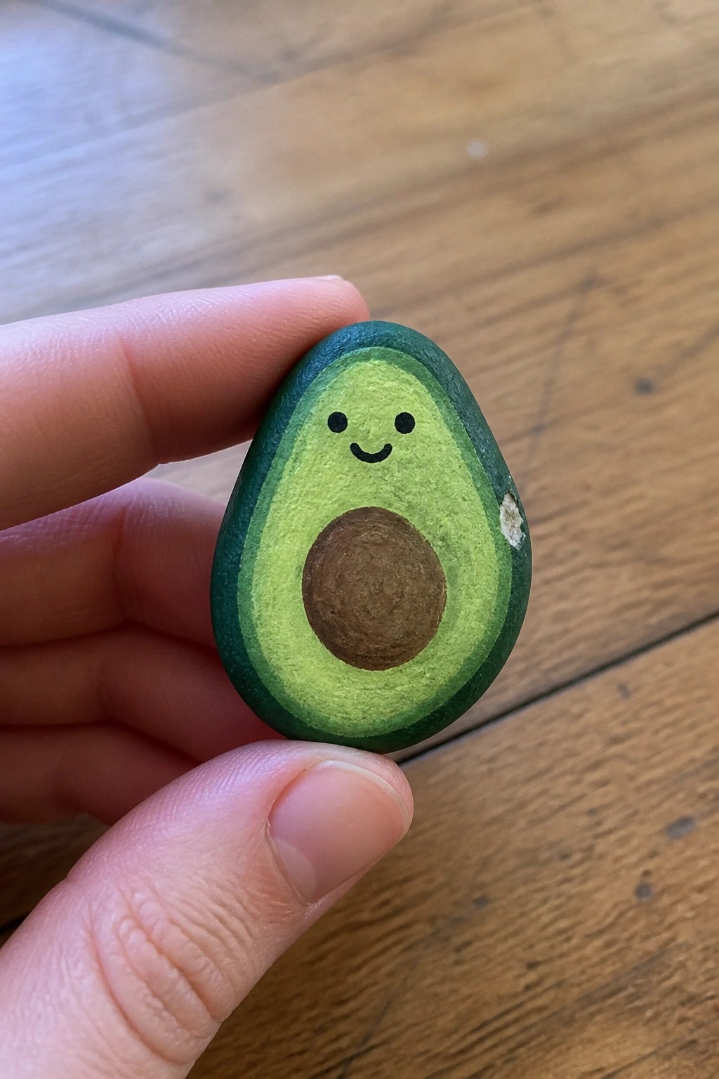 A hand holds a small oval rock painted to look like an avocado half with green layers, a brown pit, and a simple smiling face.