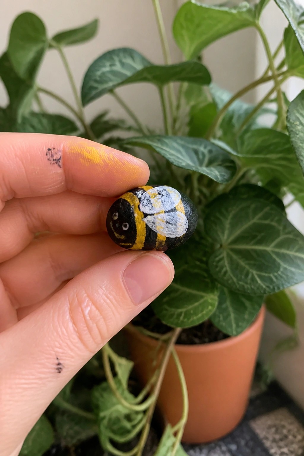 Hand holding black-yellow bumblebee painted rock, paint smudges on fingers, plants behind.