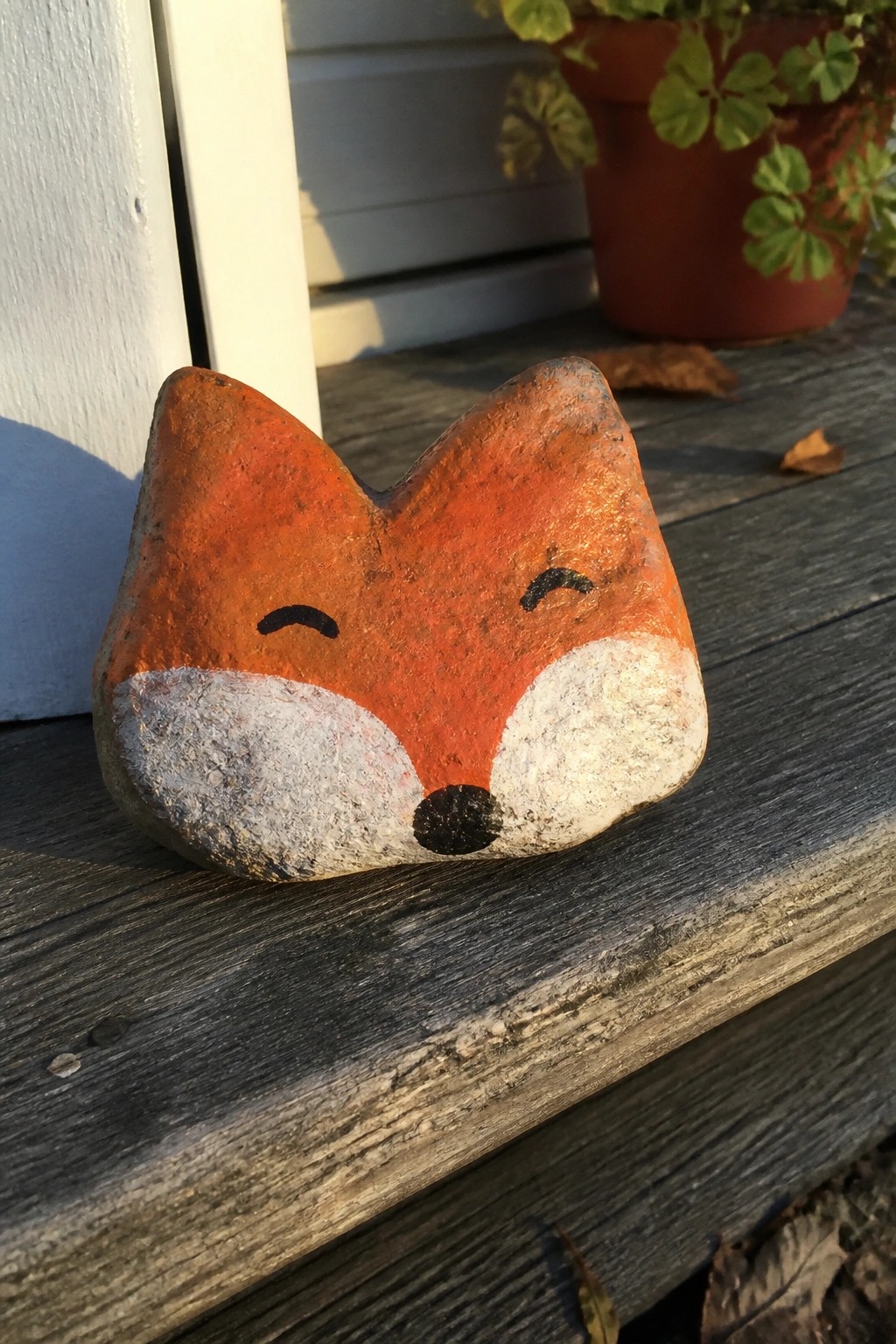 A painted rock with an orange fox face, white cheeks, and black nose and eyes sits on a wooden surface.