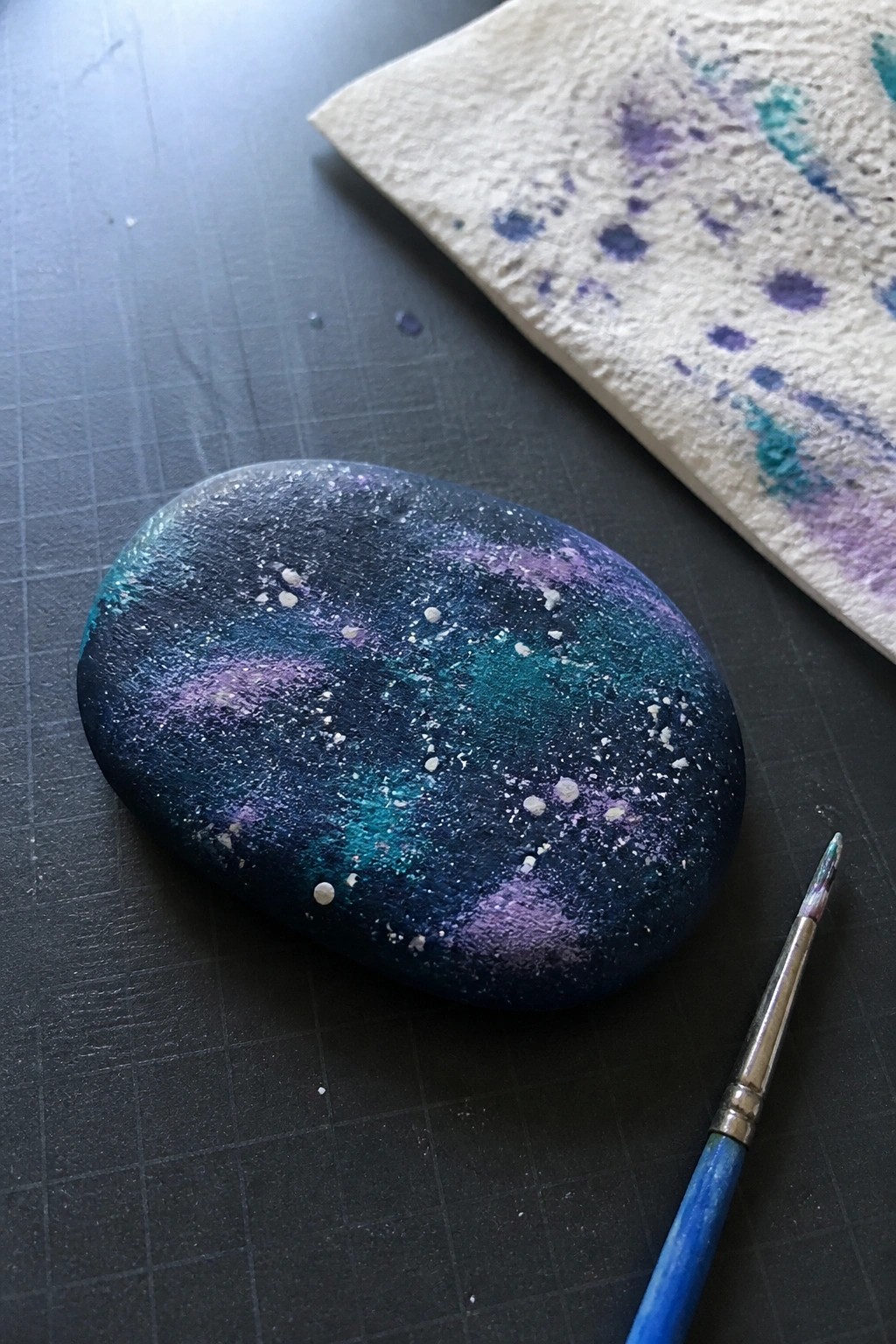 A smooth oval rock painted with blended blue, teal, and purple colors and scattered white dots sits on a dark surface beside a blue paintbrush and a paint-stained paper towel.