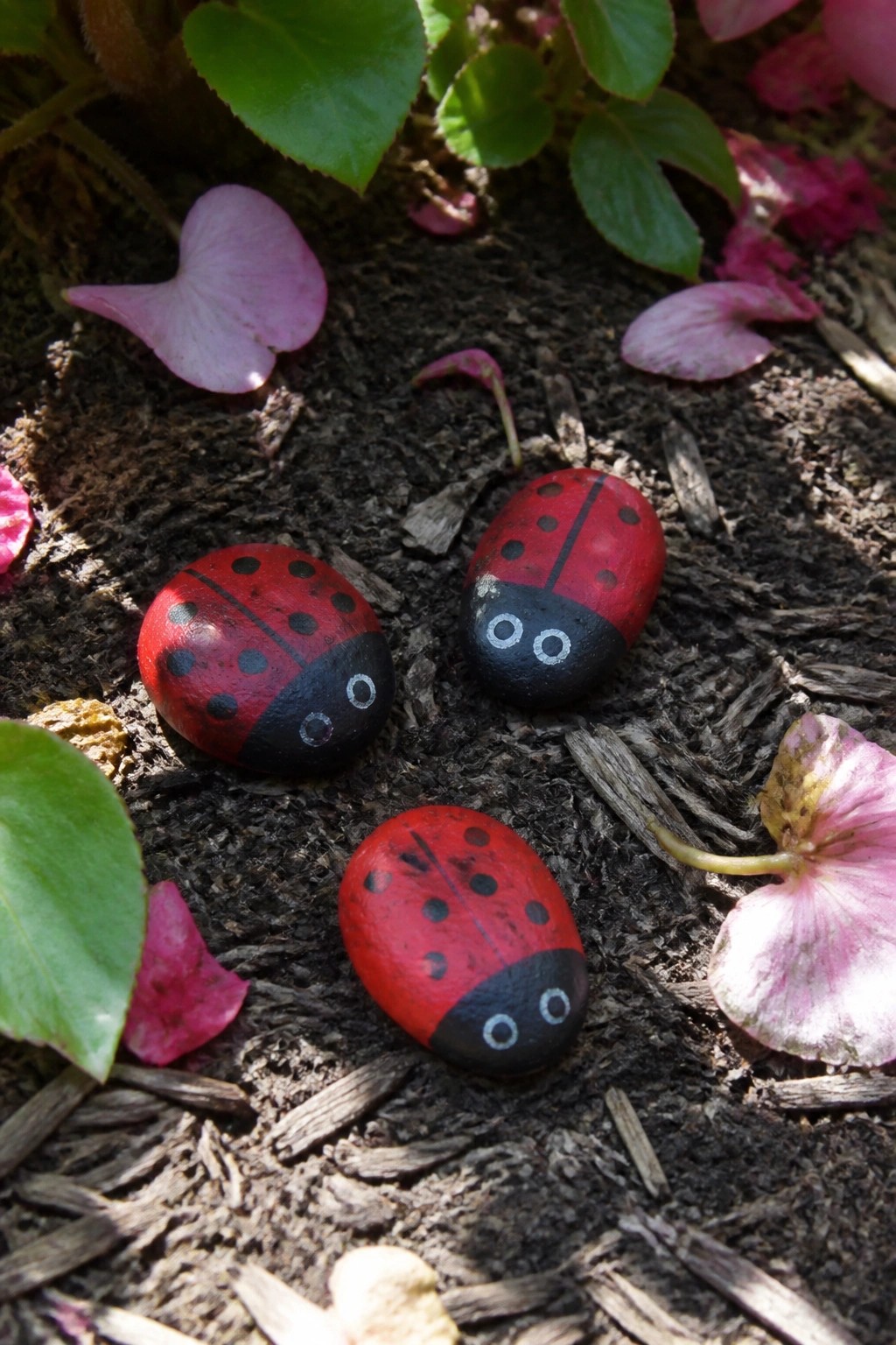 Three red ladybug rocks with black spots on soil amid pink petals and leaves.