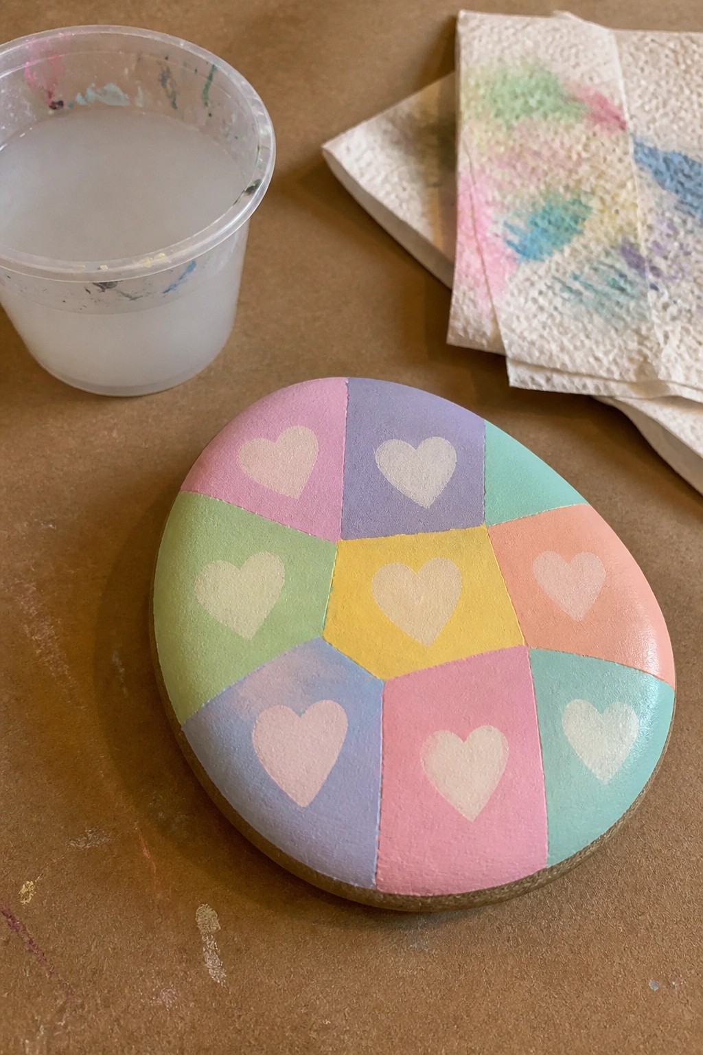 Painted rock with pastel segments and white hearts beside cup and stained towels