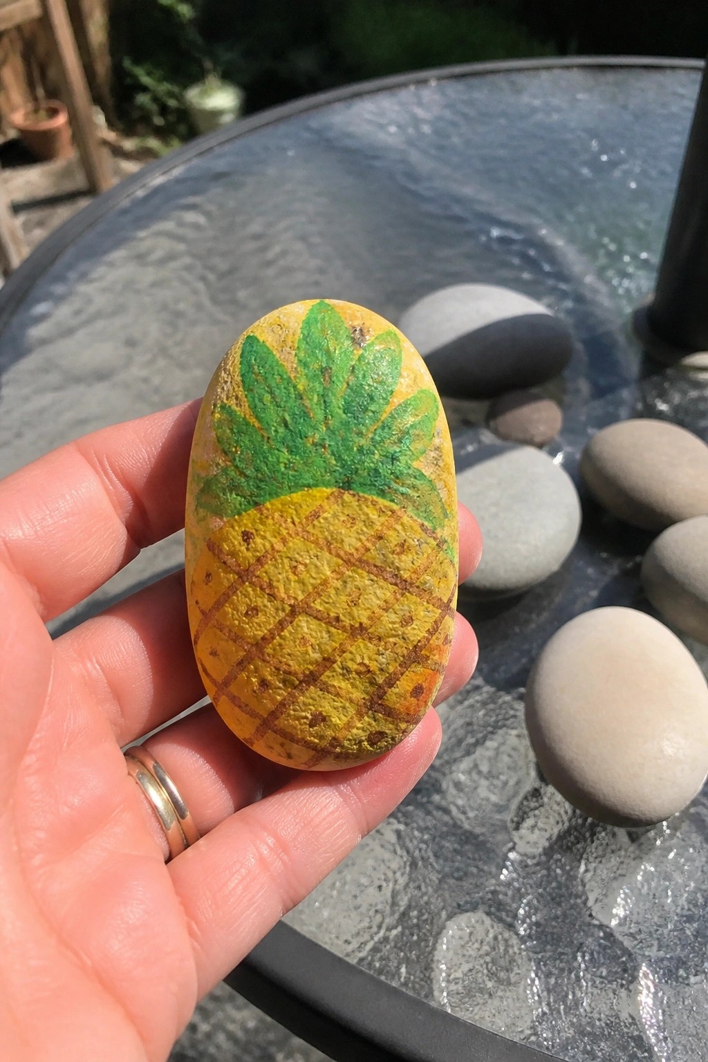 Hand holding yellow pineapple-painted rock above glass table with stones