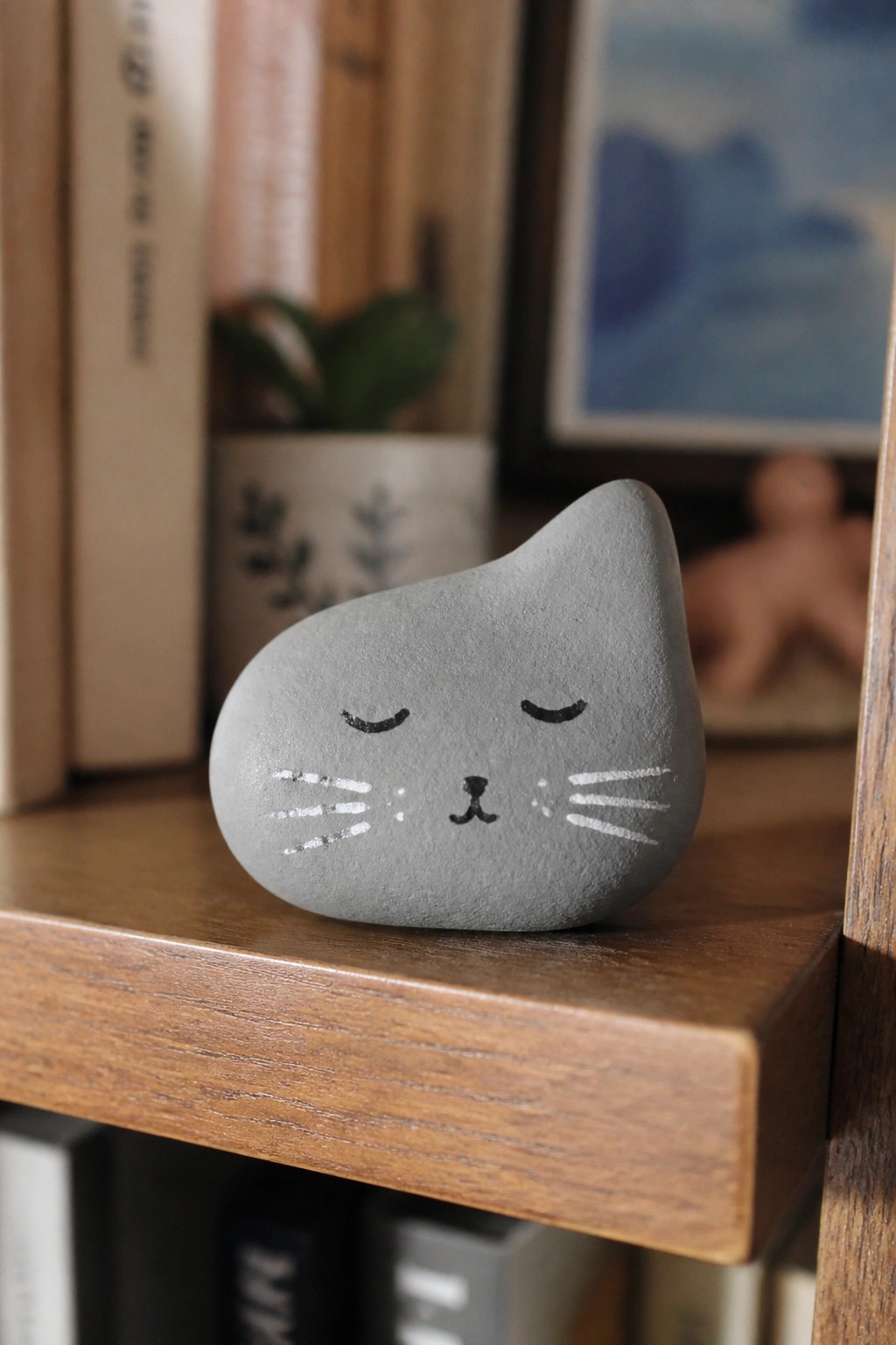 Gray cat stone with painted face resting on wooden shelf edge.