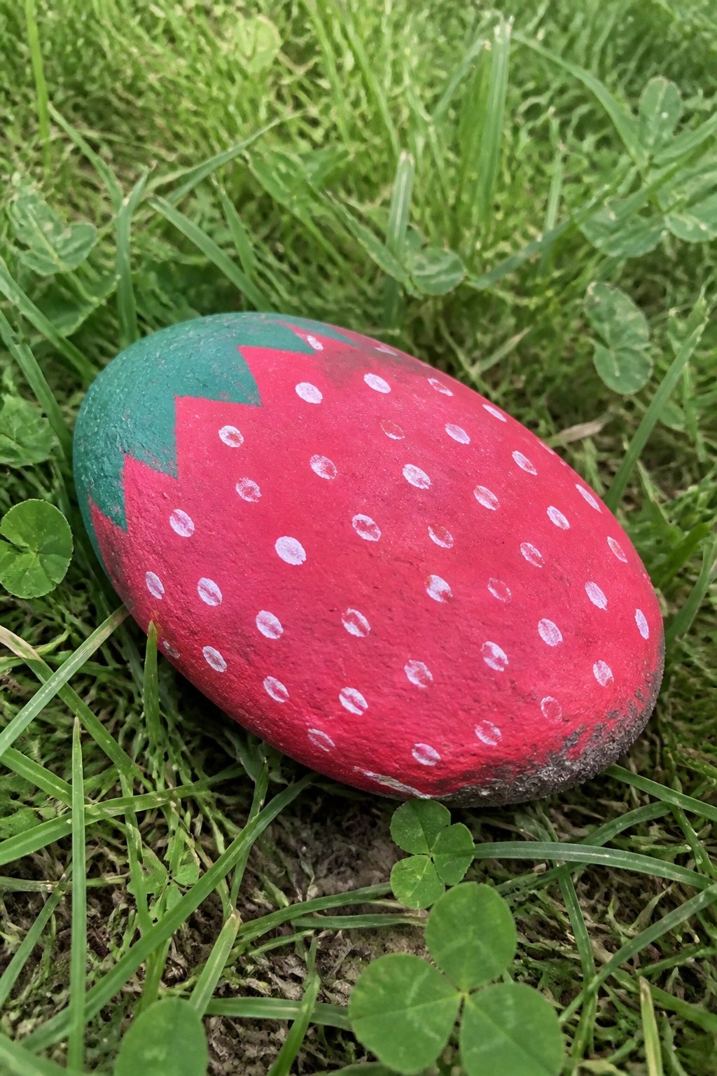 Painted rock resembling strawberry with red body, white dots, green top on grass
