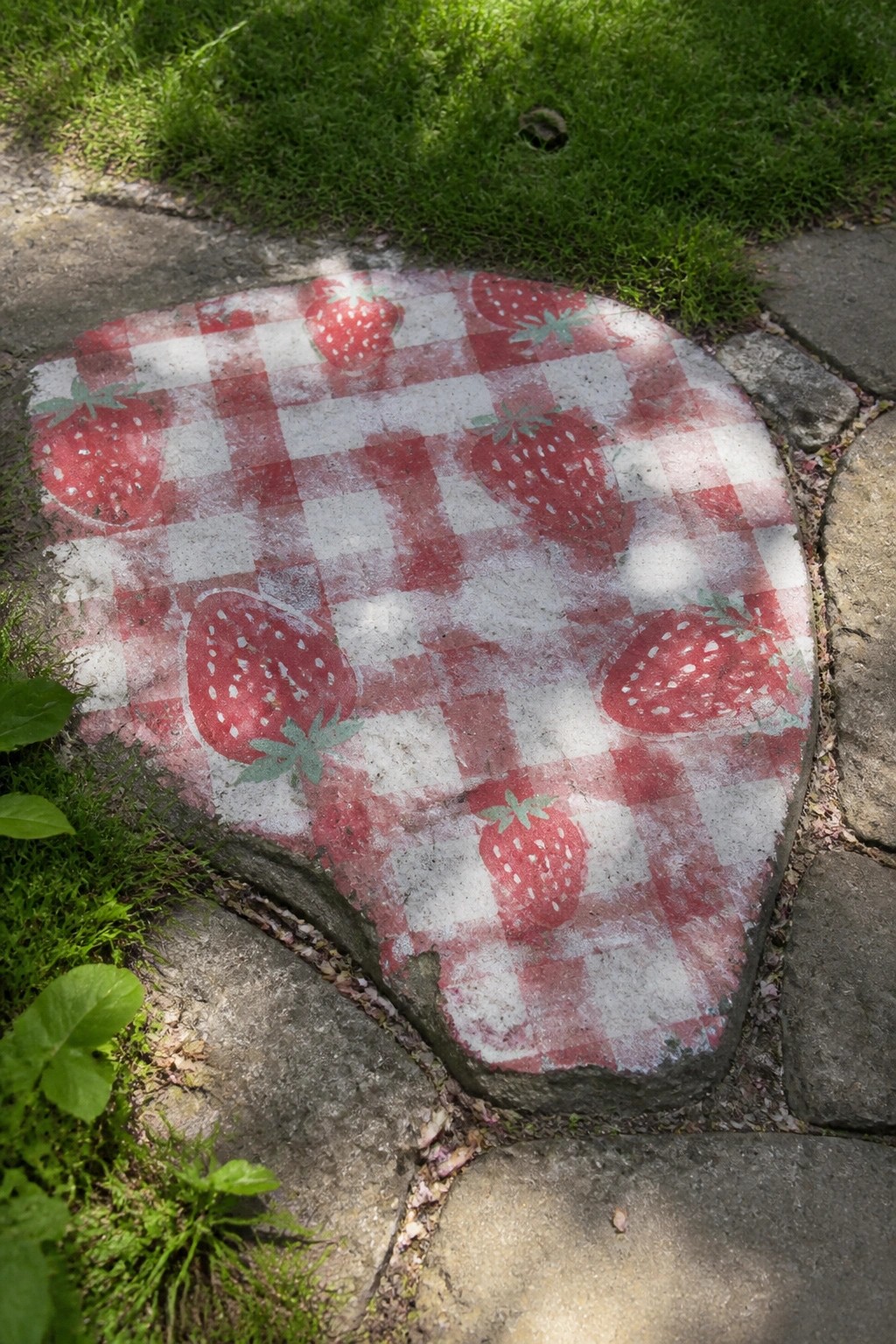 A large irregular rock painted with a red and white checkered pattern and several strawberries rests on a stone path next to grass.
