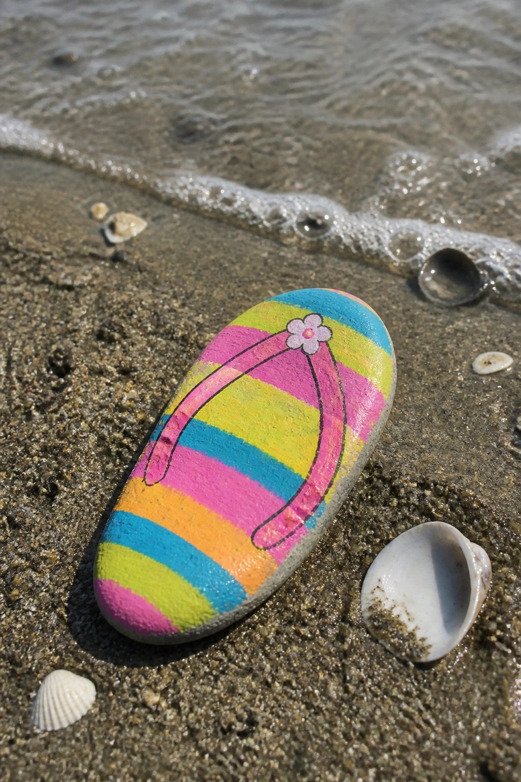 A smooth oval rock painted with colorful horizontal stripes to look like a flip-flop, resting on wet sand near the water with shells around it.