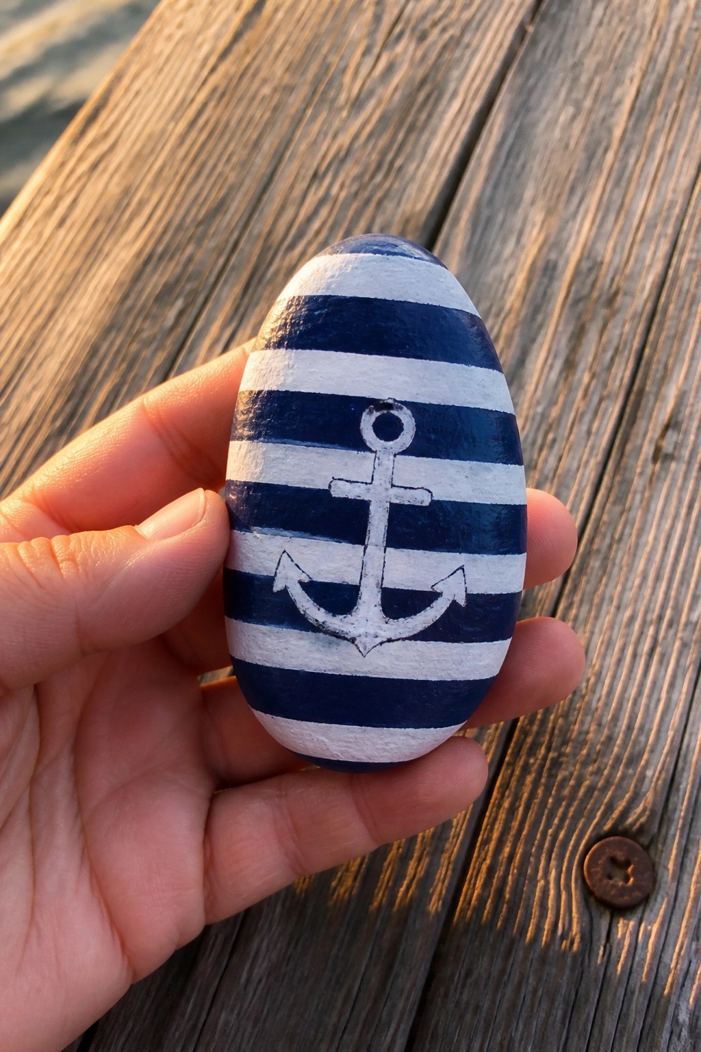Hand holding blue-and-white striped rock with anchor on wooden deck.