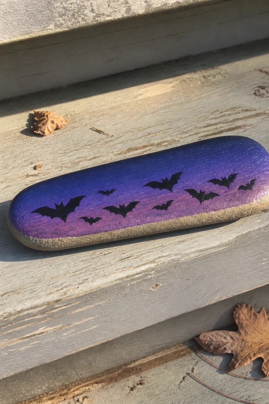 An elongated painted rock with a blue-to-purple gradient background and black bat silhouettes scattered across the surface.