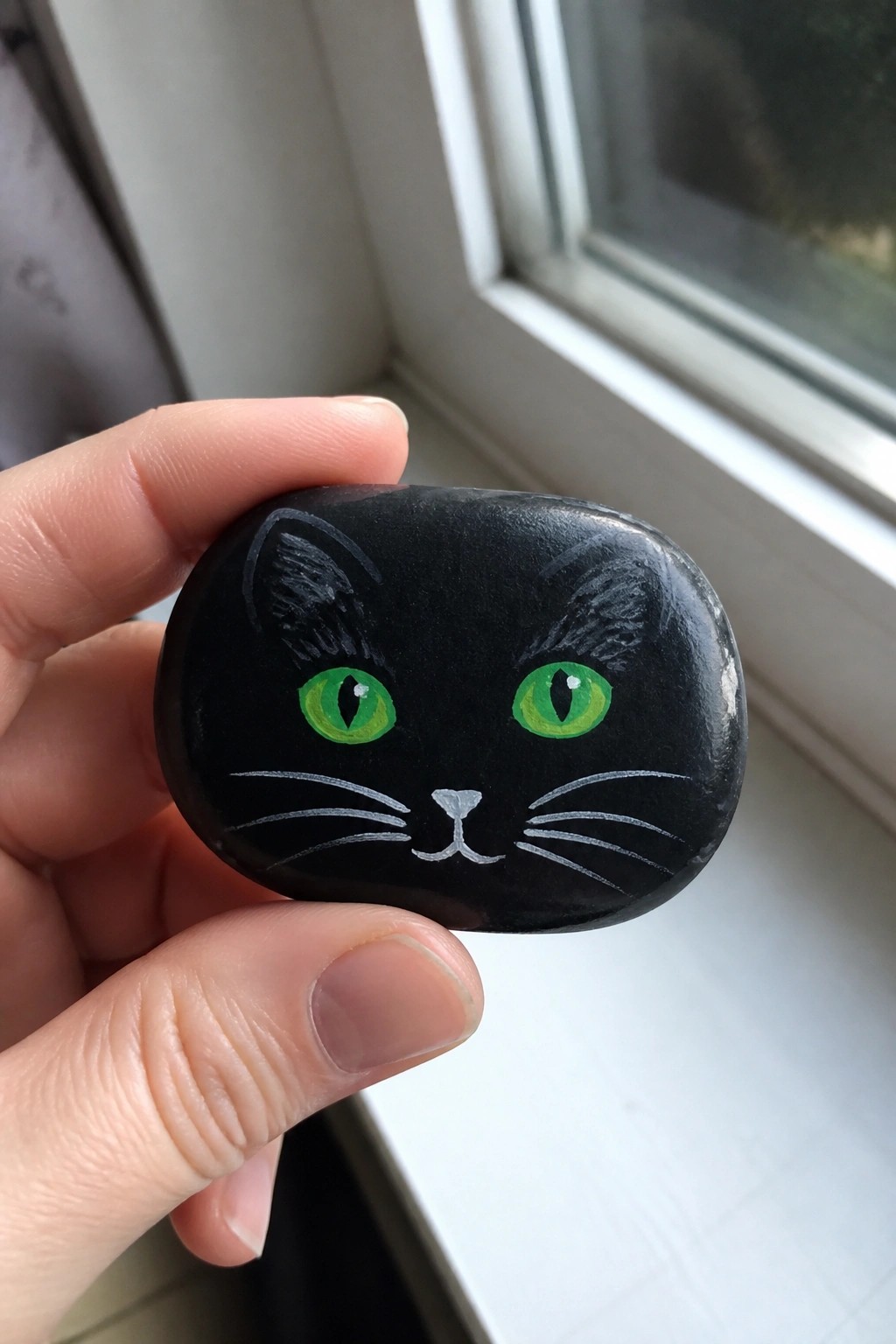 Hand holding black painted rock with green-eyed cat face and whiskers