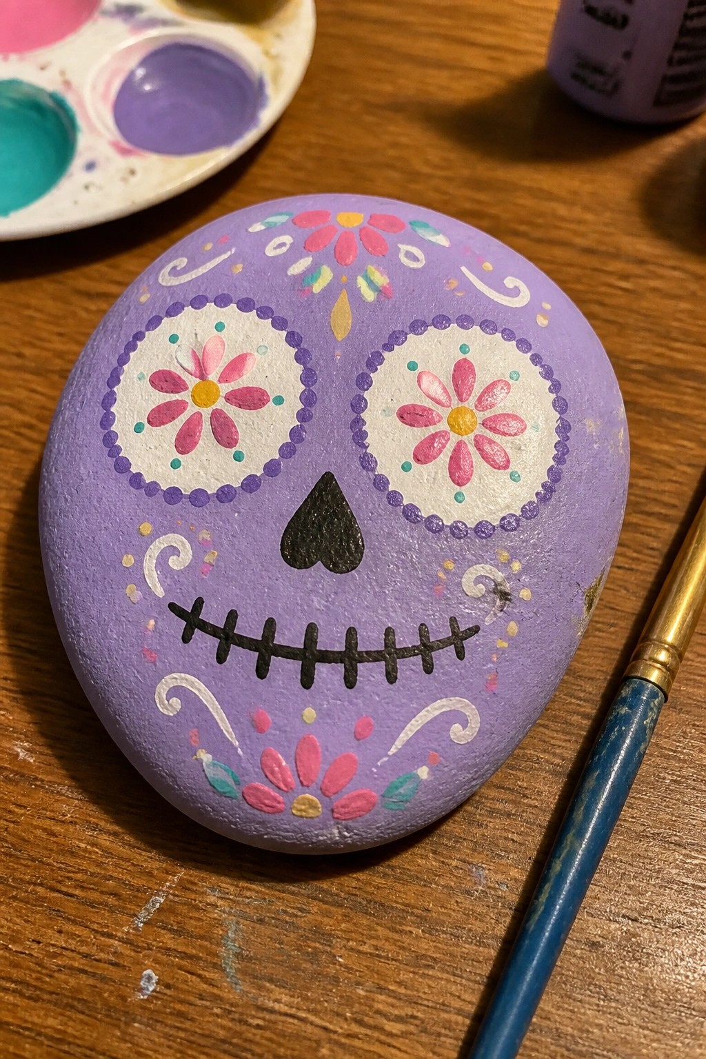 A purple painted rock decorated as a sugar skull with flower eyes, a heart nose, and a stitched mouth sits on a wooden table next to a paint palette and brush.