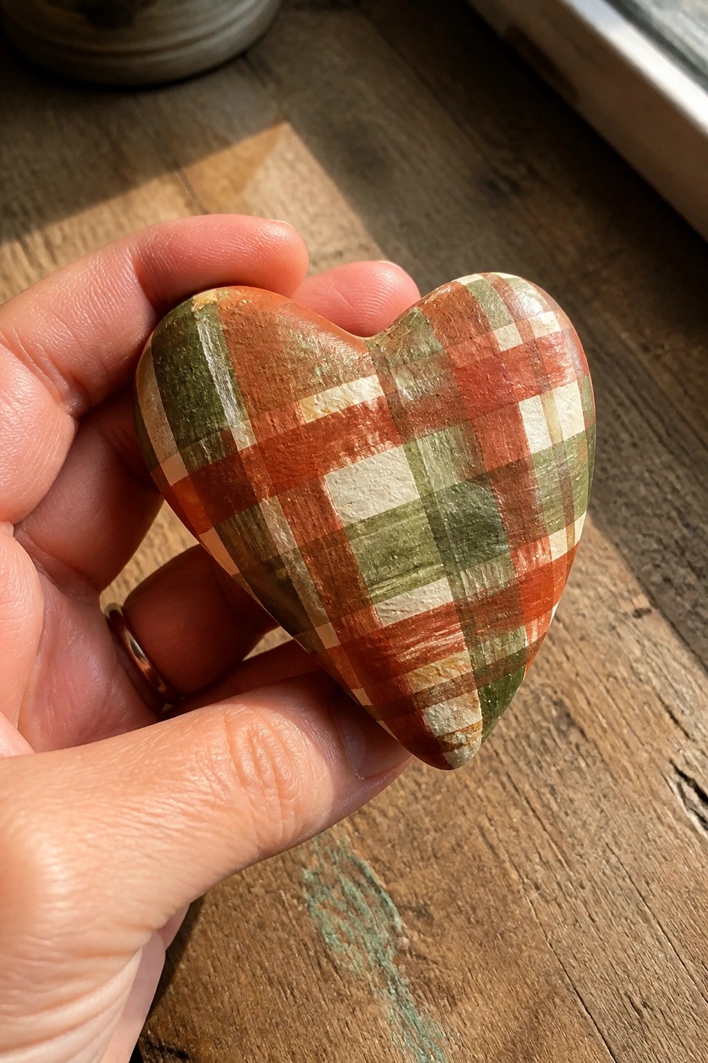 Hand holding plaid heart-shaped stone on sunlit wooden surface.