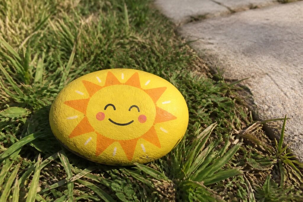 24 Uplifting Painted Rock Ideas for Garden Paths and Summer Nature Walks Yellow painted rock with smiling sun face on grass by sidewalk