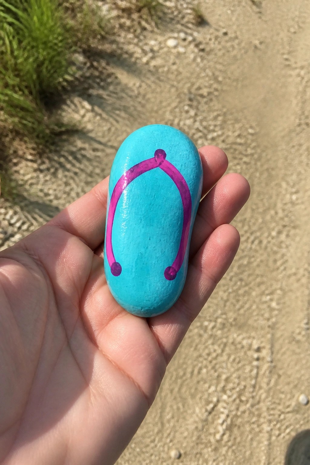 Hand holding turquoise oval rock with magenta U-shaped design on sandy path.