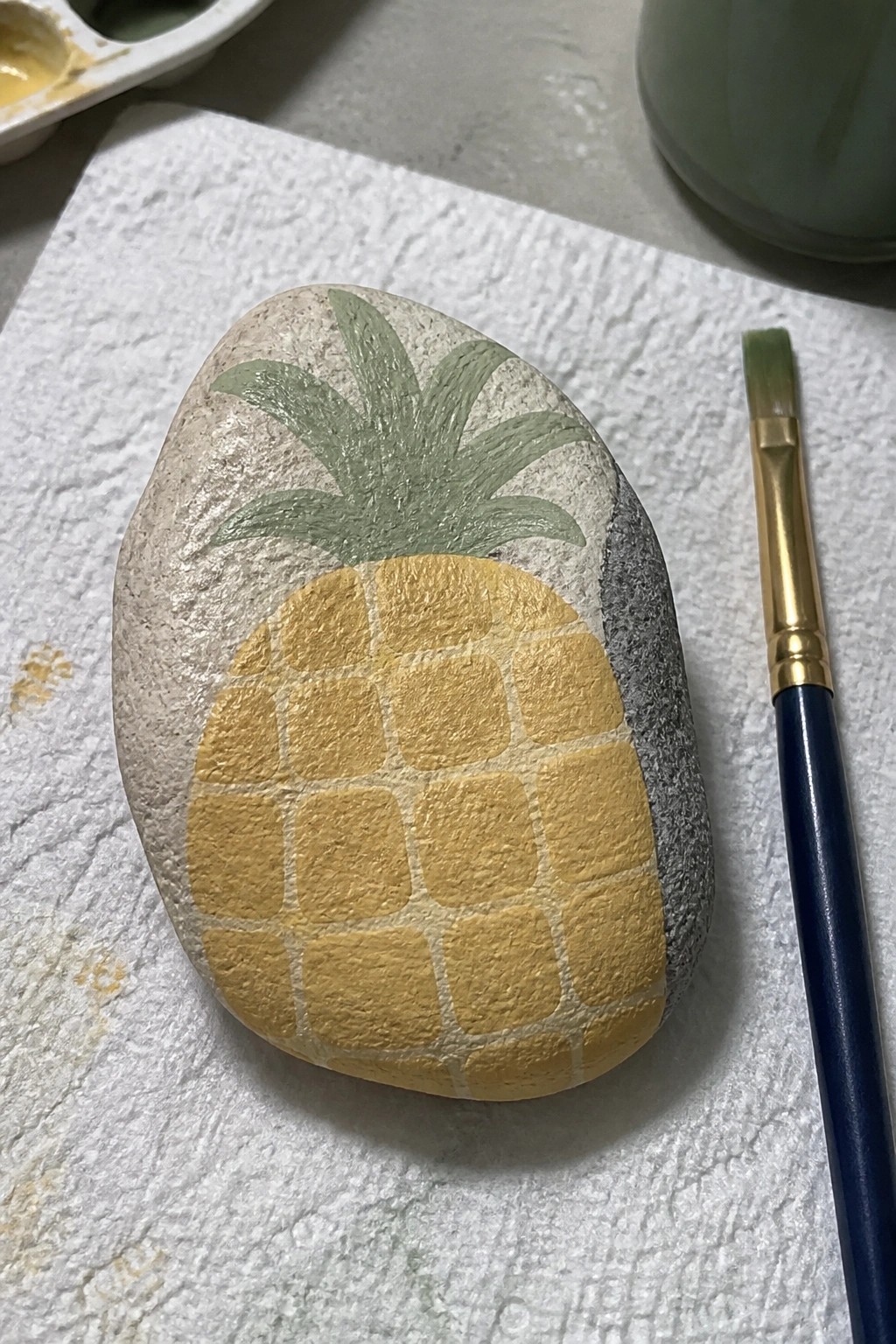 A rounded stone painted with a yellow pineapple body in a grid pattern and green leaves on top rests on a paper towel next to a paintbrush.