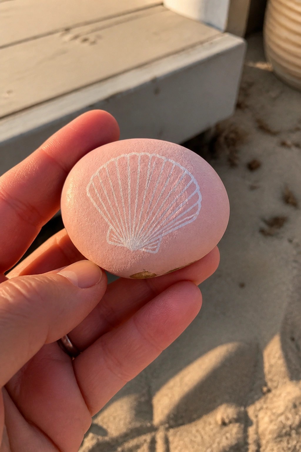 Hand holding smooth pink stone etched with white seashell on sandy ground