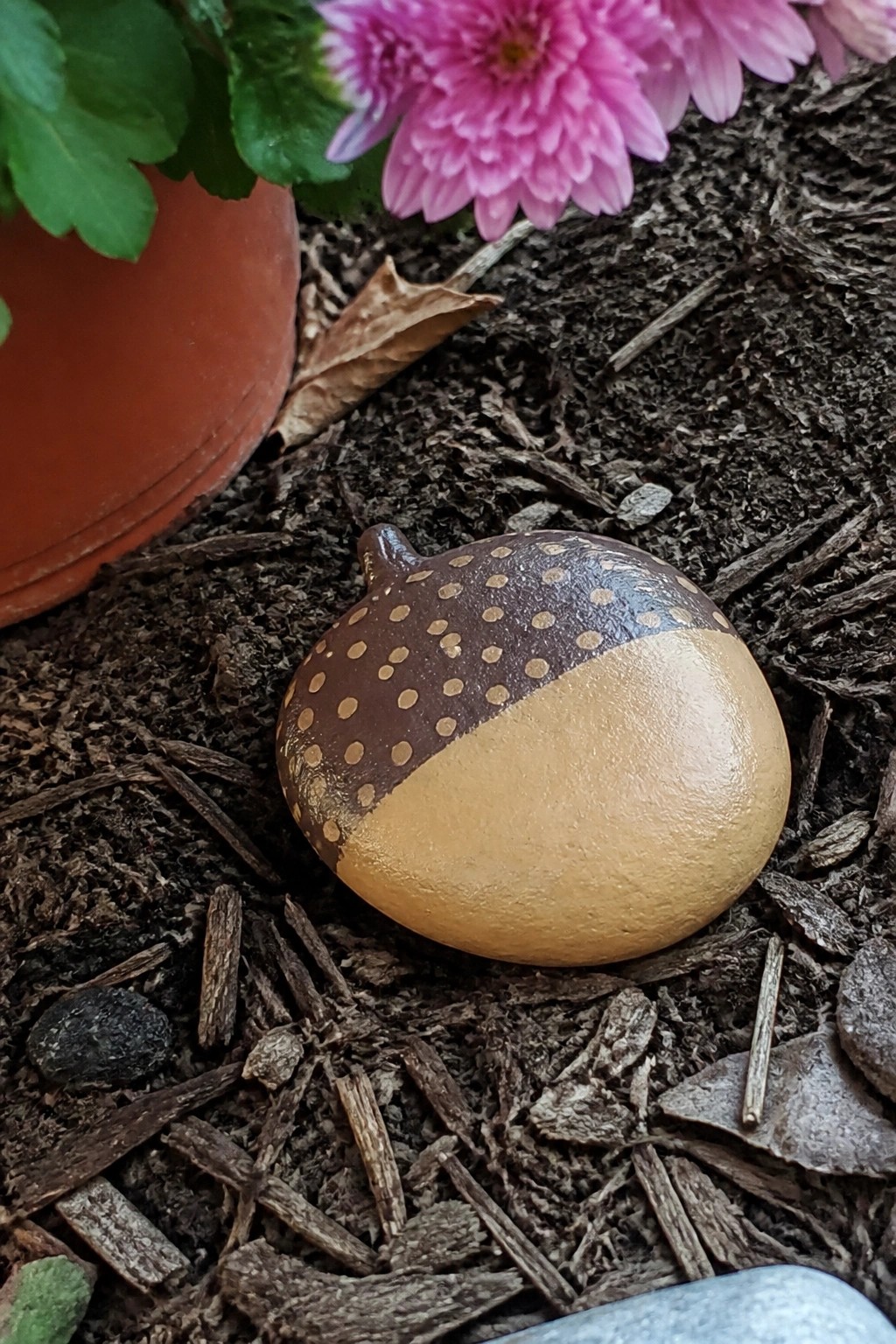 A rounded rock painted as an acorn with a dark brown dotted cap and tan body sits on dark mulch beside a terracotta pot and pink flower.