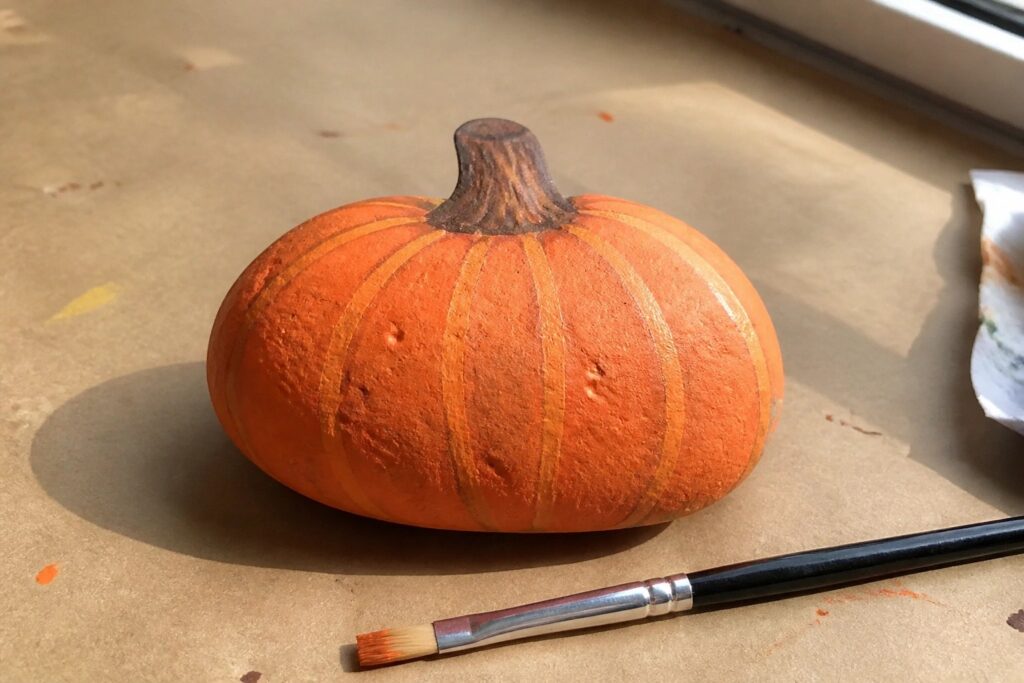 25 Beginner-Friendly Fall Painted Rock Ideas for Weekend DIY Crafts Orange pumpkin with brown stem beside blue paintbrush on brown surface