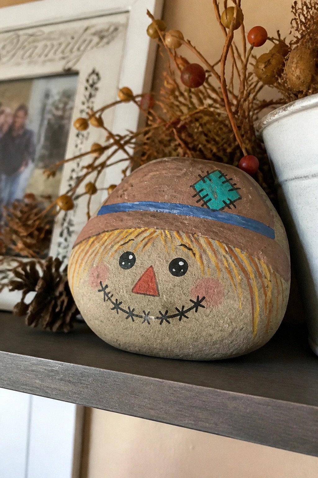 Painted scarecrow rock with brown hat, blue band, stitched face on shelf