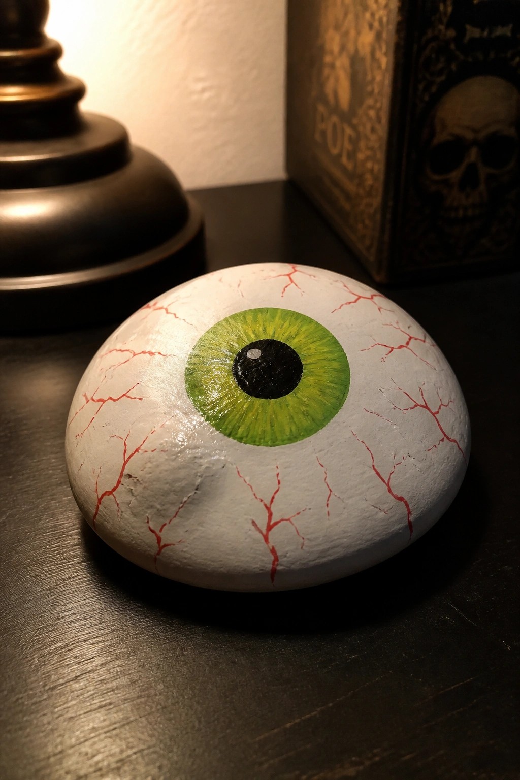 A smooth oval rock painted as a green eyeball with red veins rests on a dark surface next to a lamp base and book.