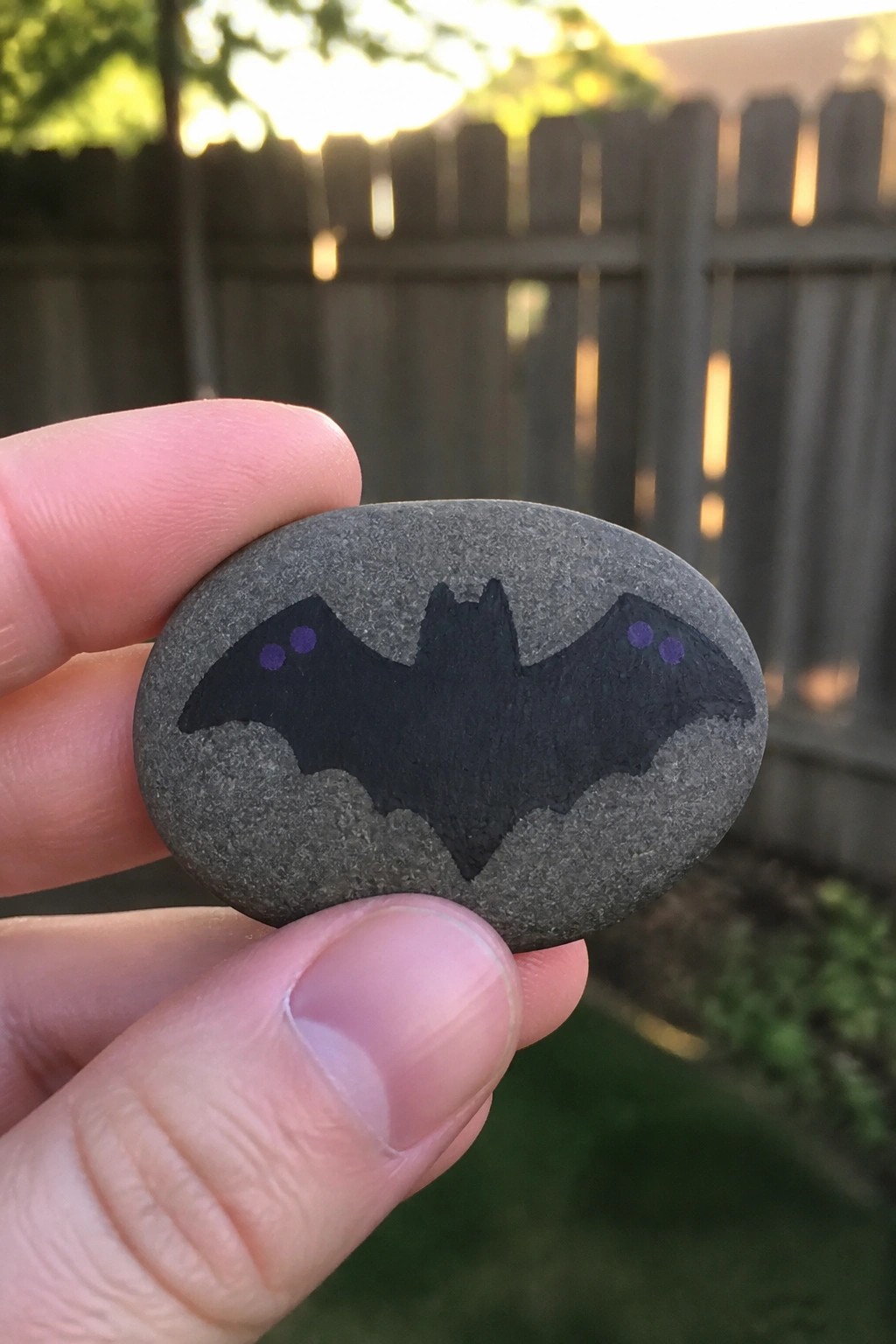 Hand holding gray stone painted with black bat and purple eyes