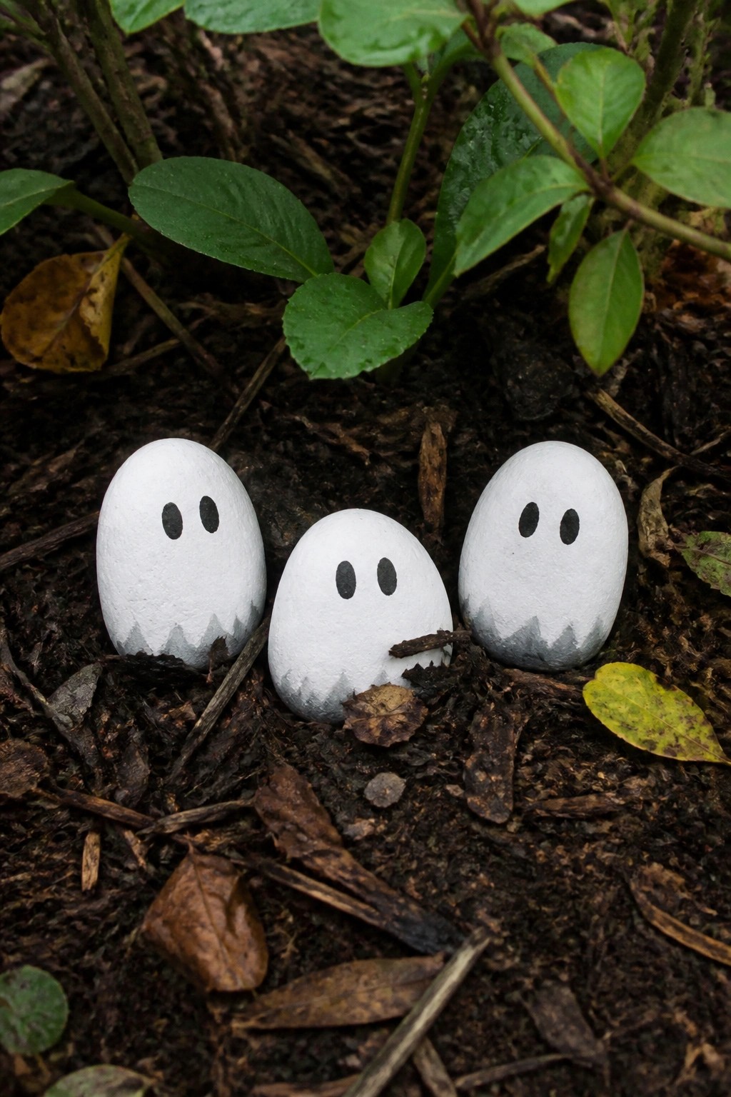 Three white ghost rocks with black eyes on leafy forest soil.